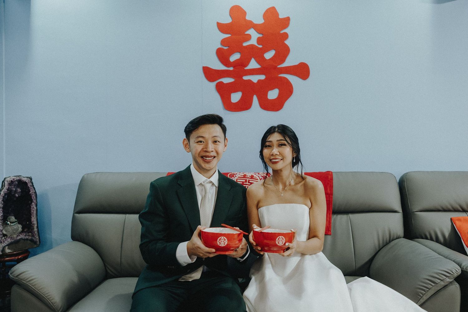 Traditional Chinese wedding ceremony with couple holding red gift boxes seated on leather couch under double happiness symbol.