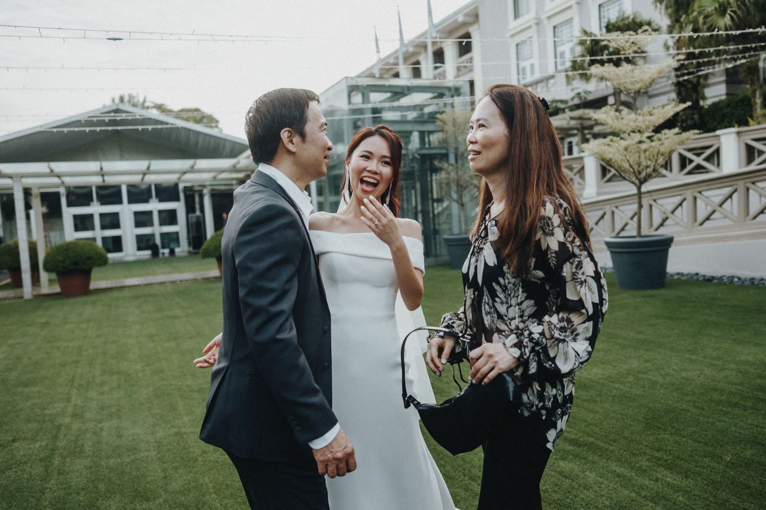 A candid moment at a wedding venue with guests laughing and interacting on a green lawn near white buildings.