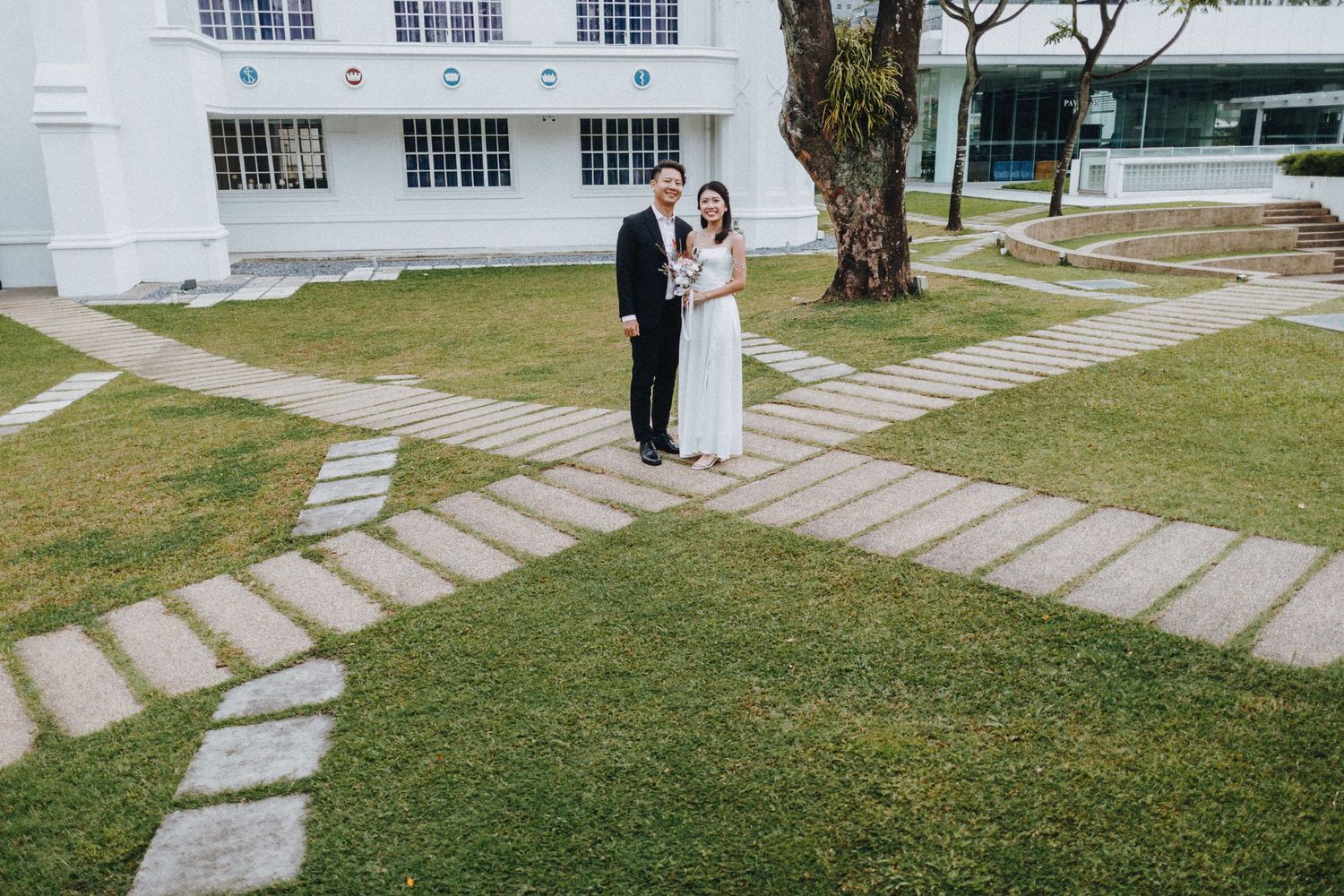 A wedding couple stands together on a stone path crossing a green lawn in front of a white building.