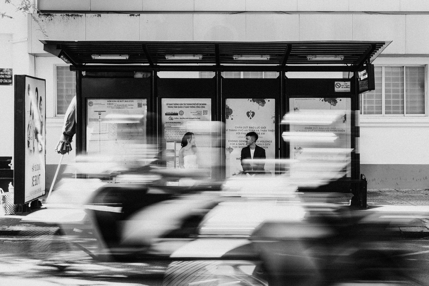 Black and white motion blur of vehicles passing a bus stop shelter with silhouettes visible inside.