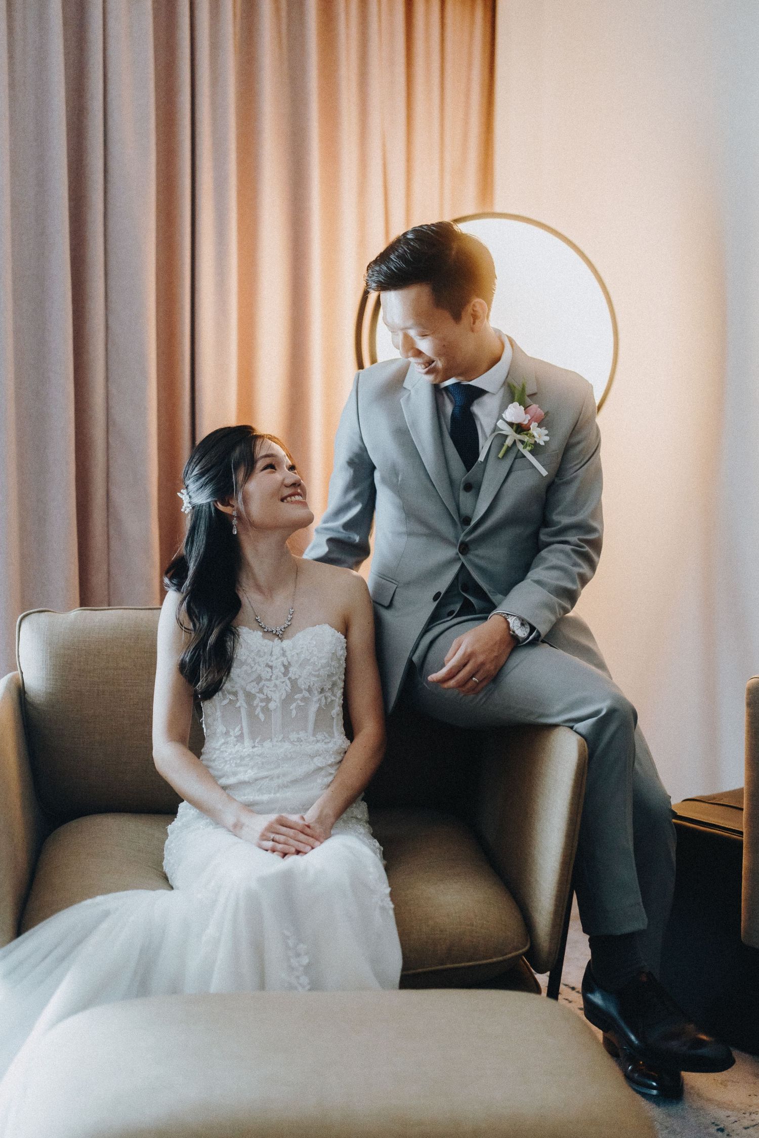 Elegant wedding portrait of couple in grey suit and white dress seated on beige armchair.