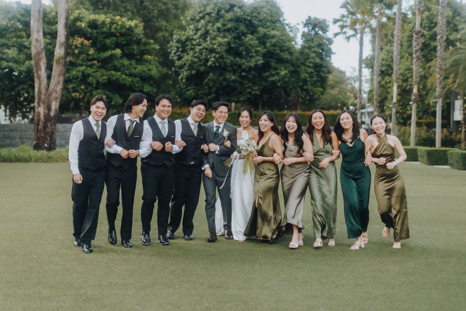 Wedding party in formal attire poses together on a green lawn during outdoor celebration.