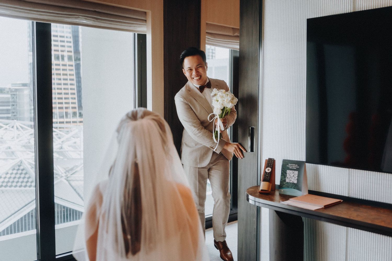 A wedding scene in a modern hotel room with a light beige suit and wedding dress visible against city views.
