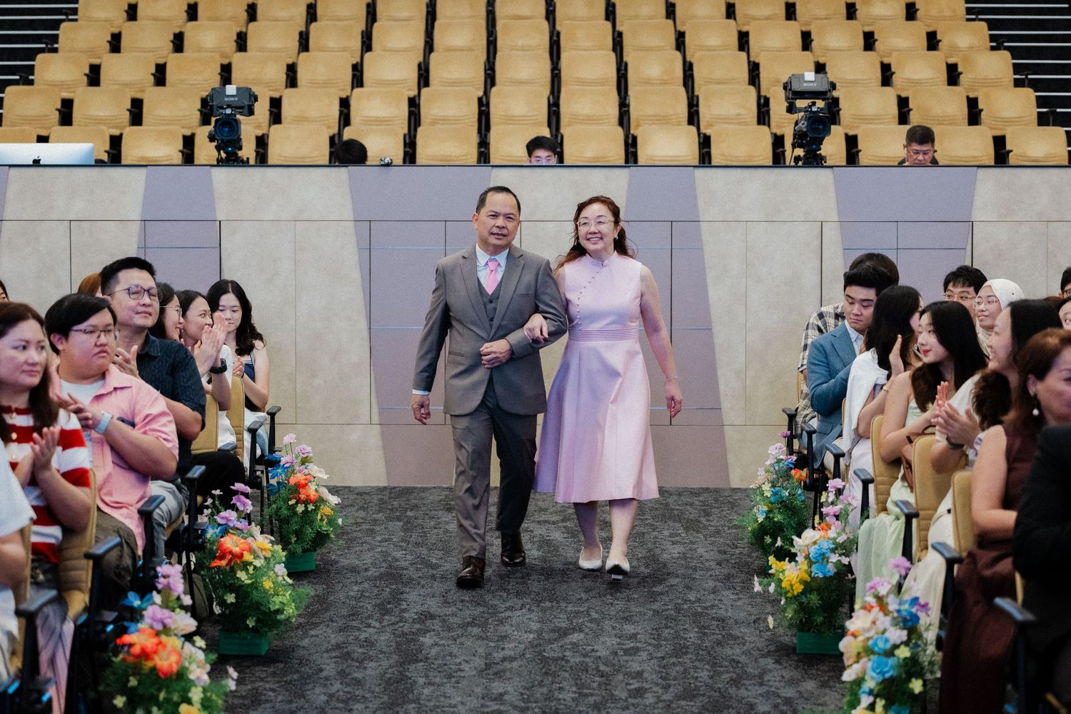 Couple walking down wedding aisle in auditorium with seated guests.