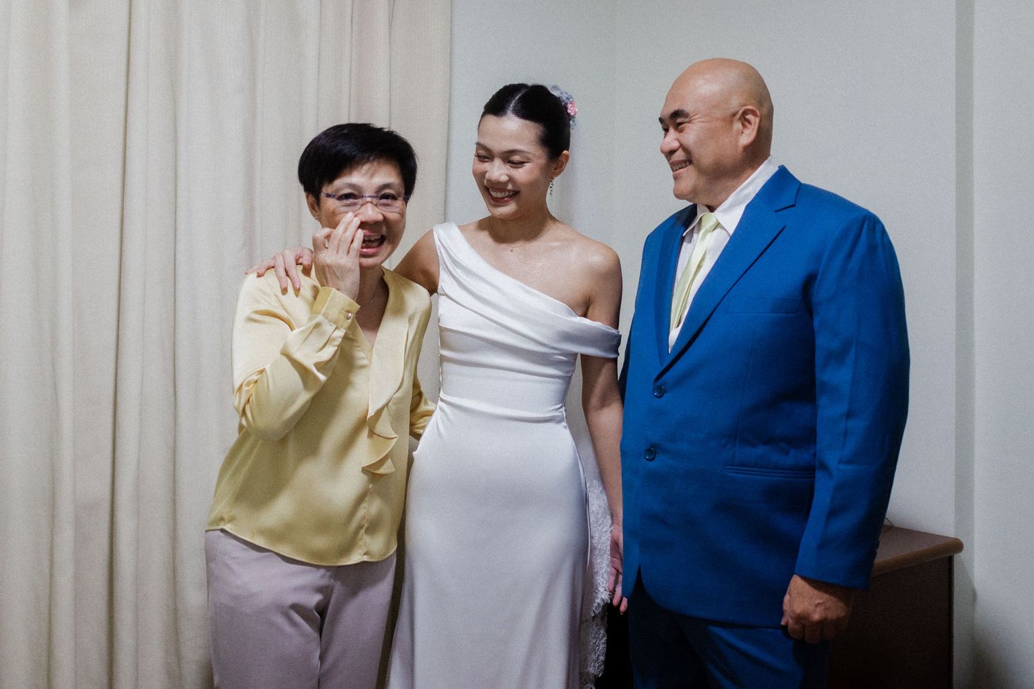 Bride in white off-shoulder gown poses with smiling older couple, woman in yellow blouse and man in blue suit.