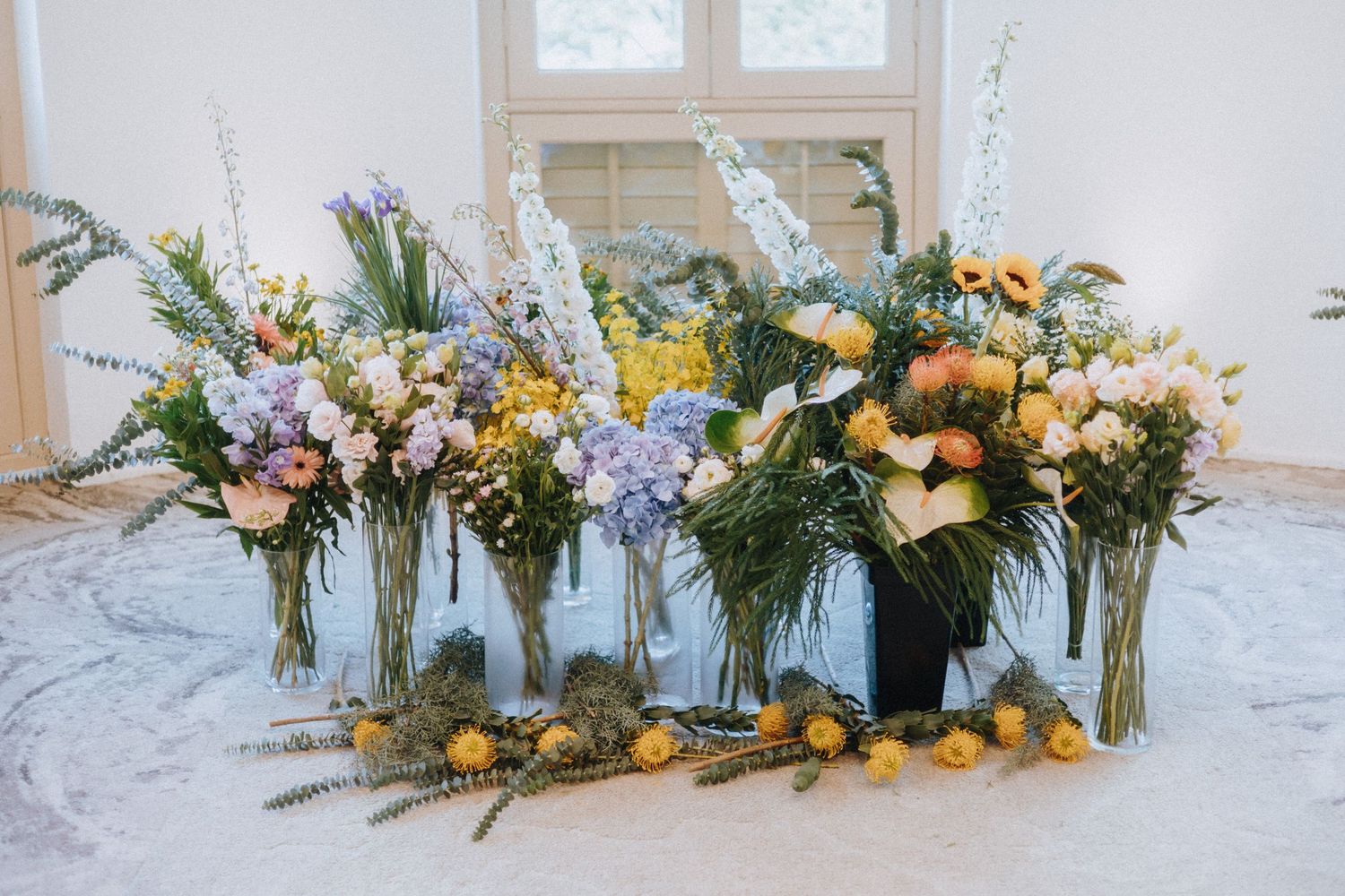 Elegant floral arrangement featuring hydrangeas, sunflowers, and wildflowers in glass vases on a white surface.