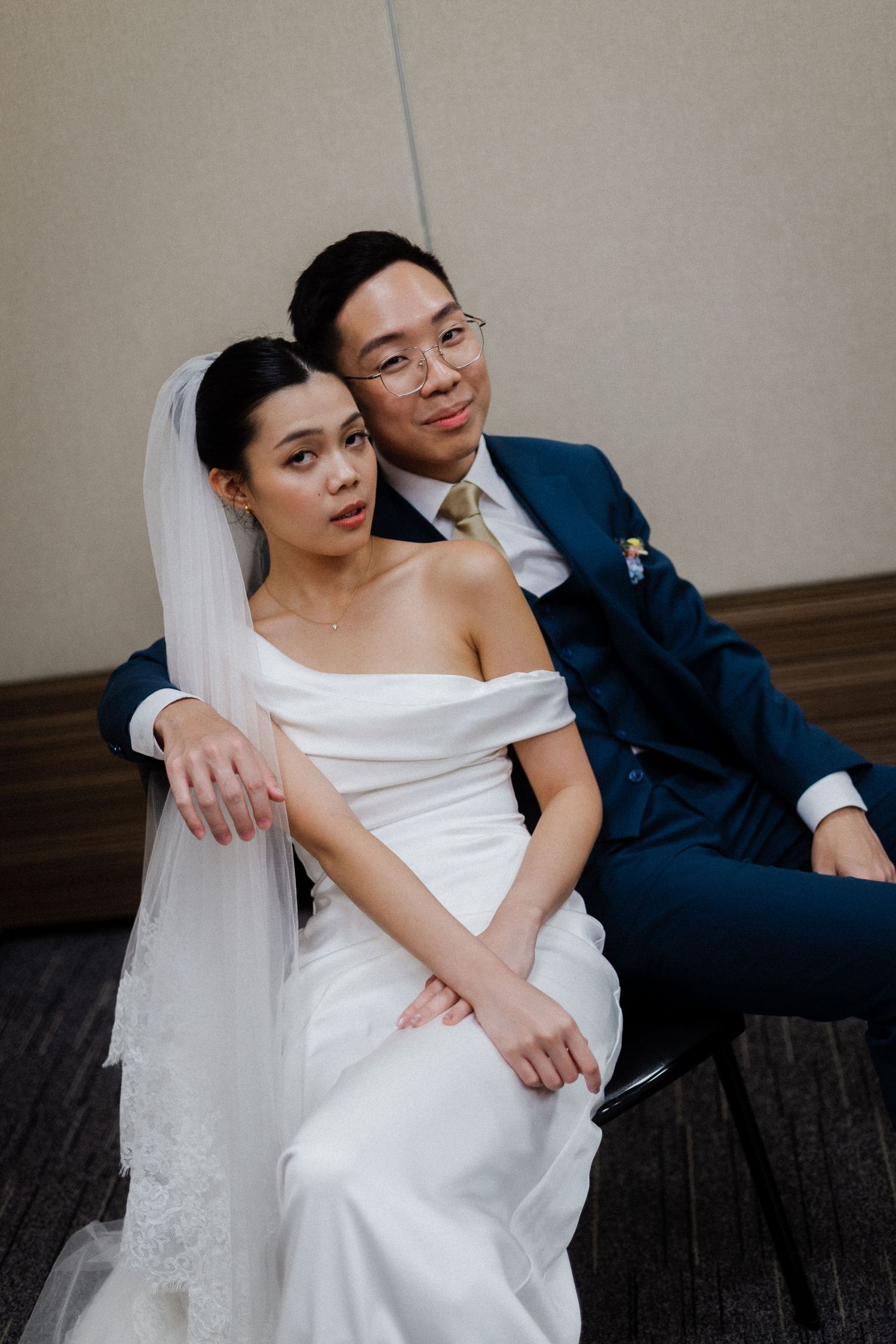 Asian bride in off-shoulder gown sits in front of groom in navy suit as they pose together intimately.