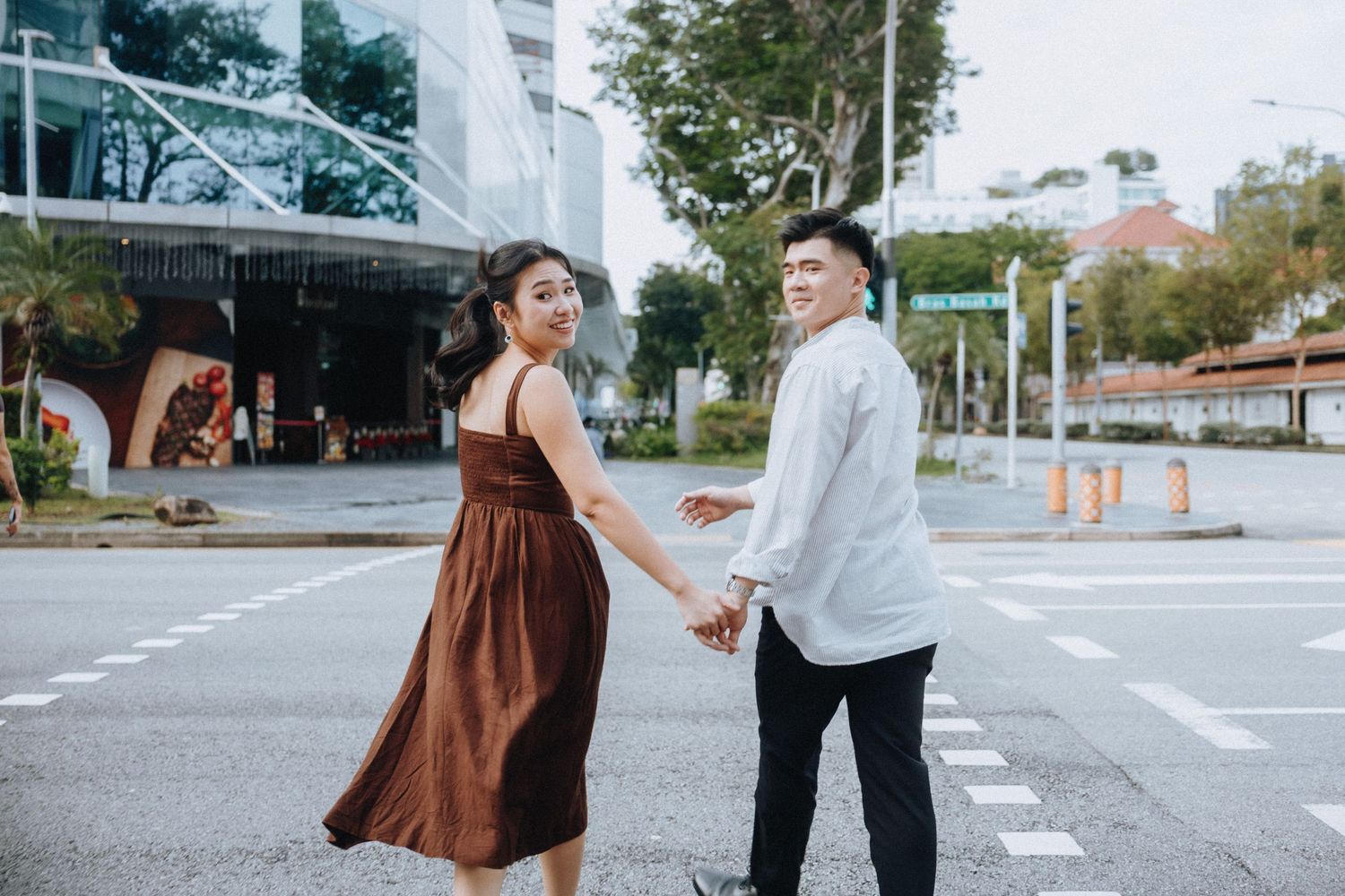 Couple holding hands while crossing a street, woman in brown dress and man in white shirt, urban background.