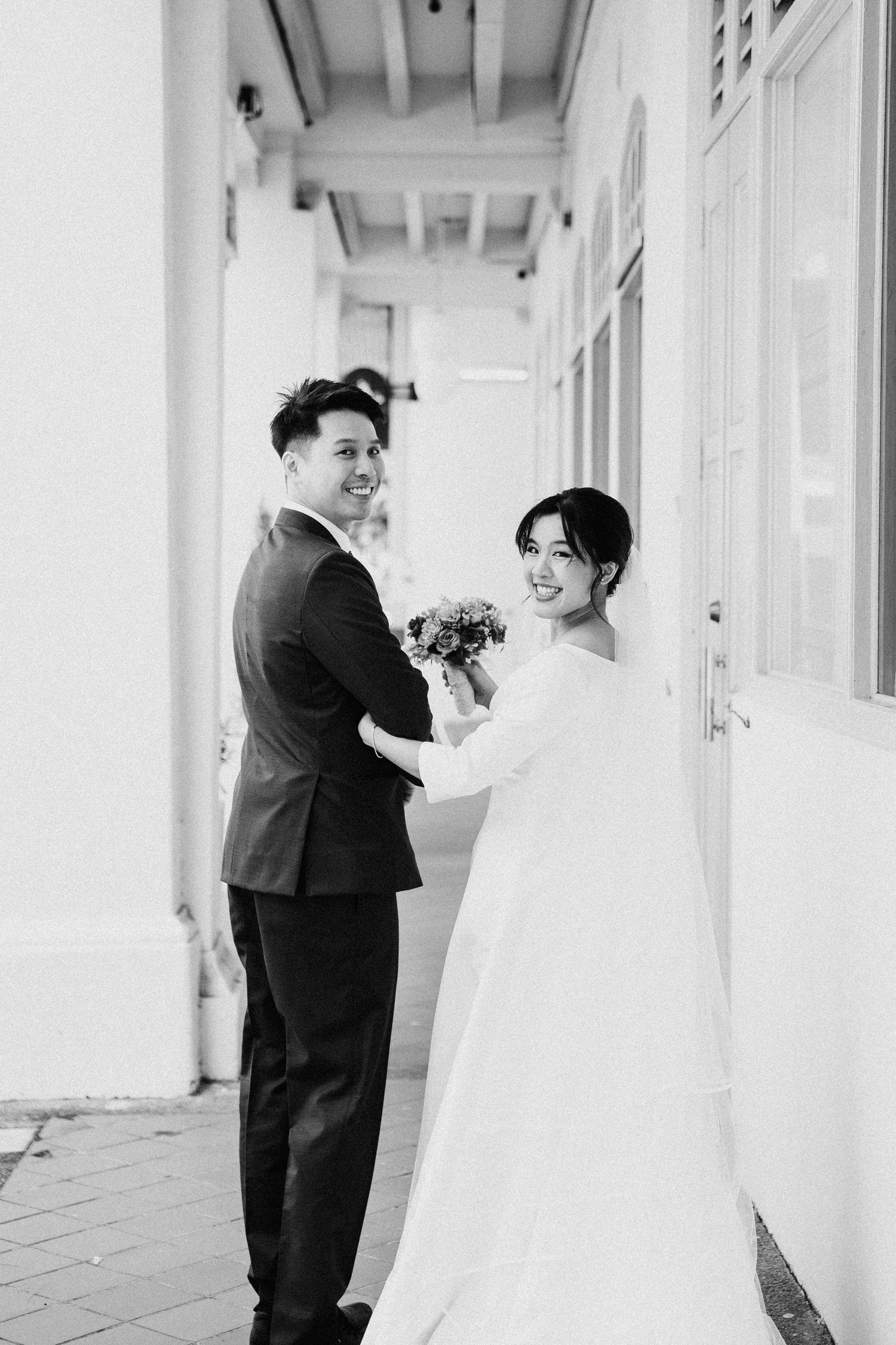 Black and white photo of smiling bride and groom posing together outside a white building on their wedding day.
