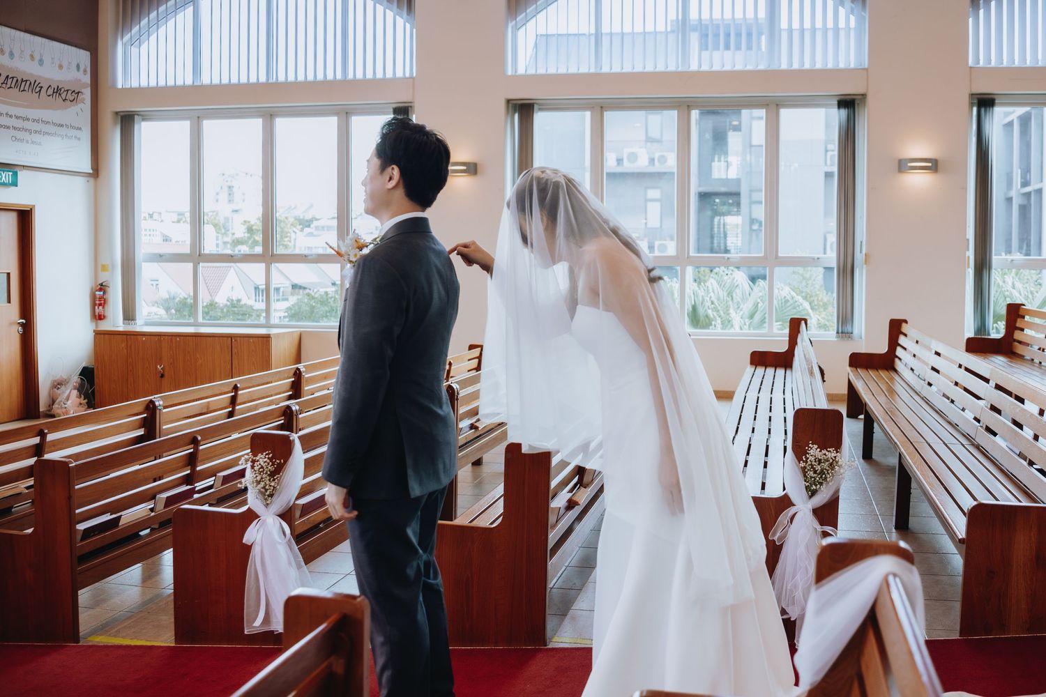 Bride and groom standing at the altar in a bright church with wooden pews decorated with white ribbons during wedding ceremony.