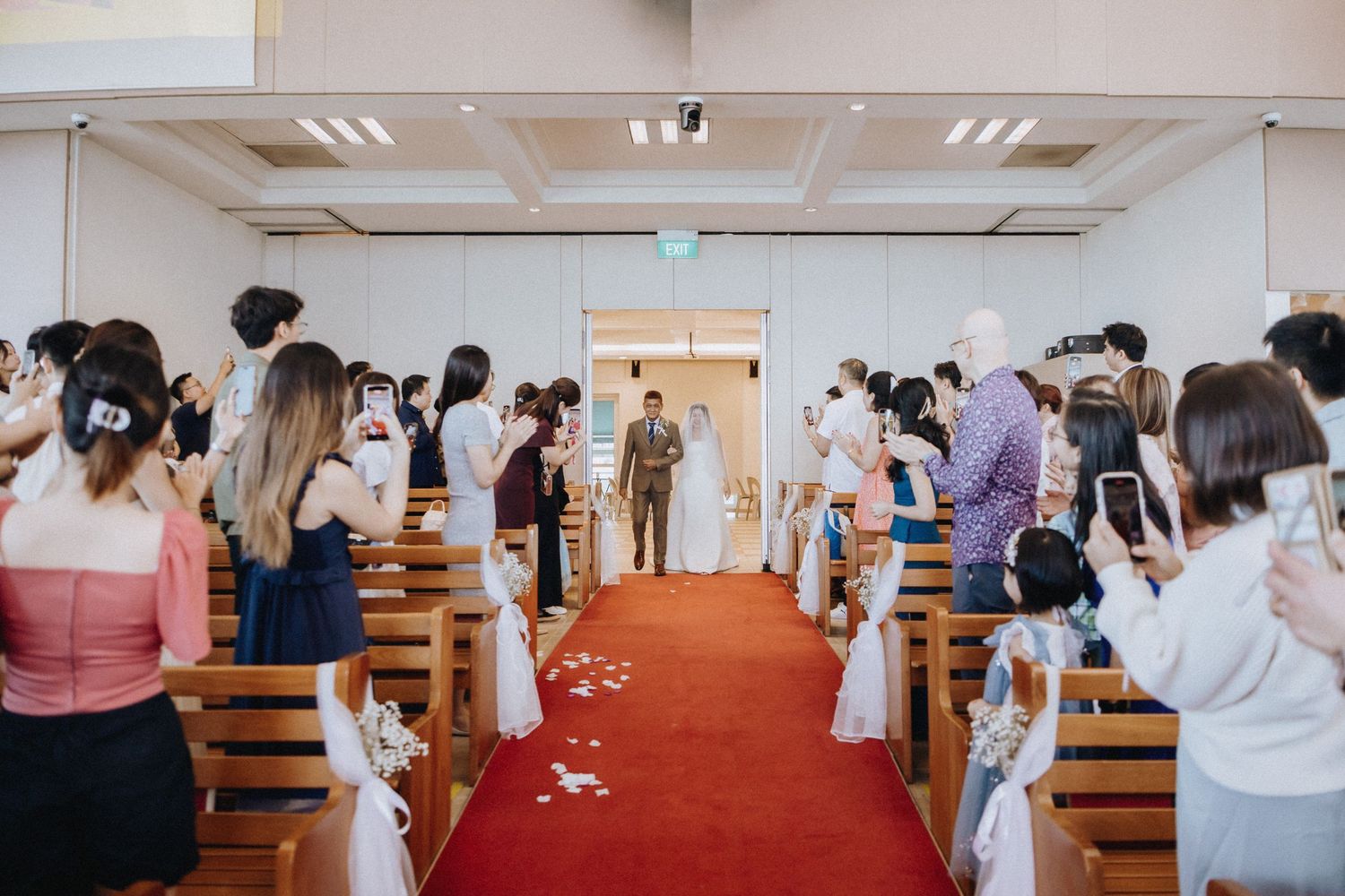 Wedding ceremony with guests seated in church pews as groom awaits bride walking down red aisle.