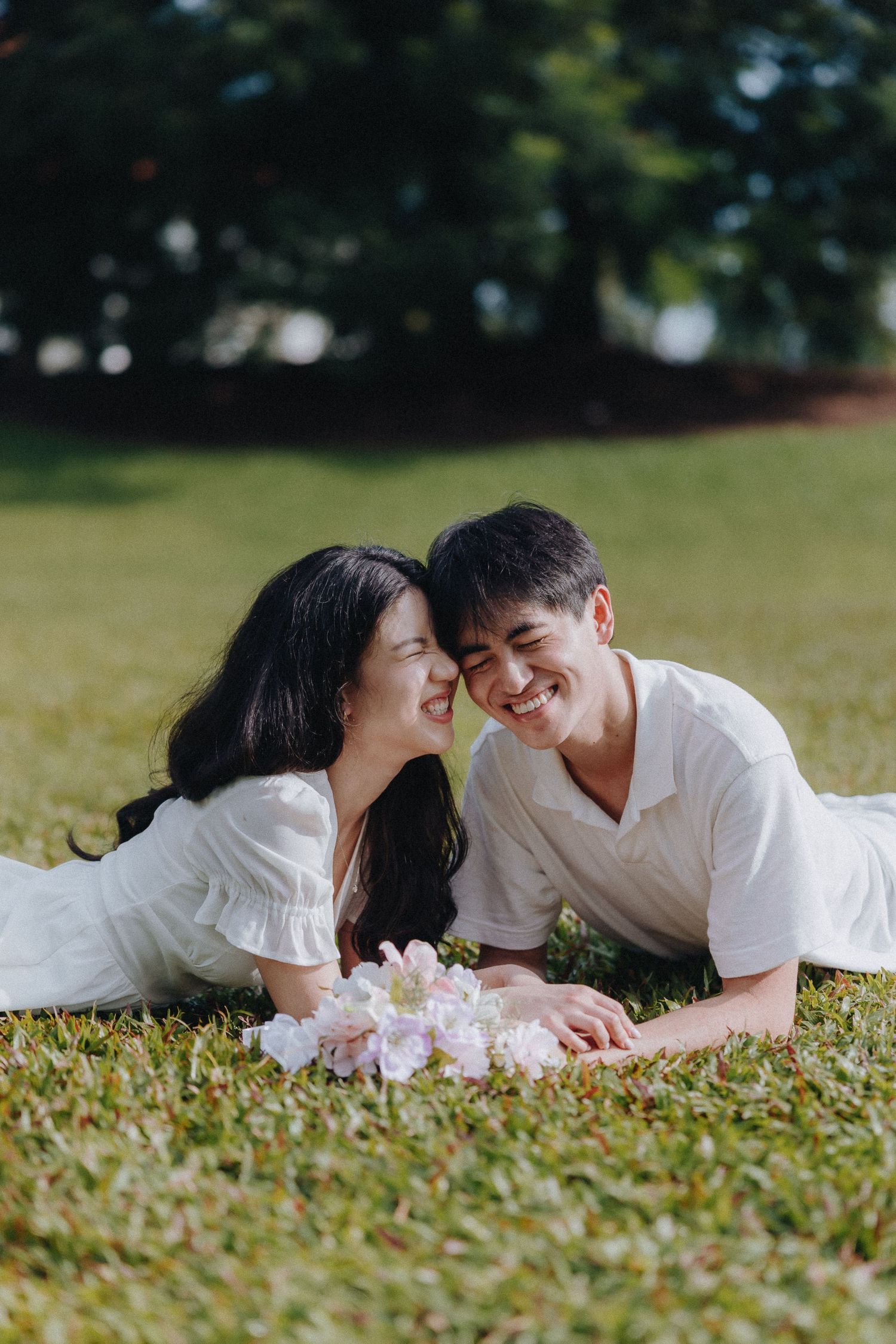 A couple lies on grass with pink flowers sharing an intimate moment outdoors.