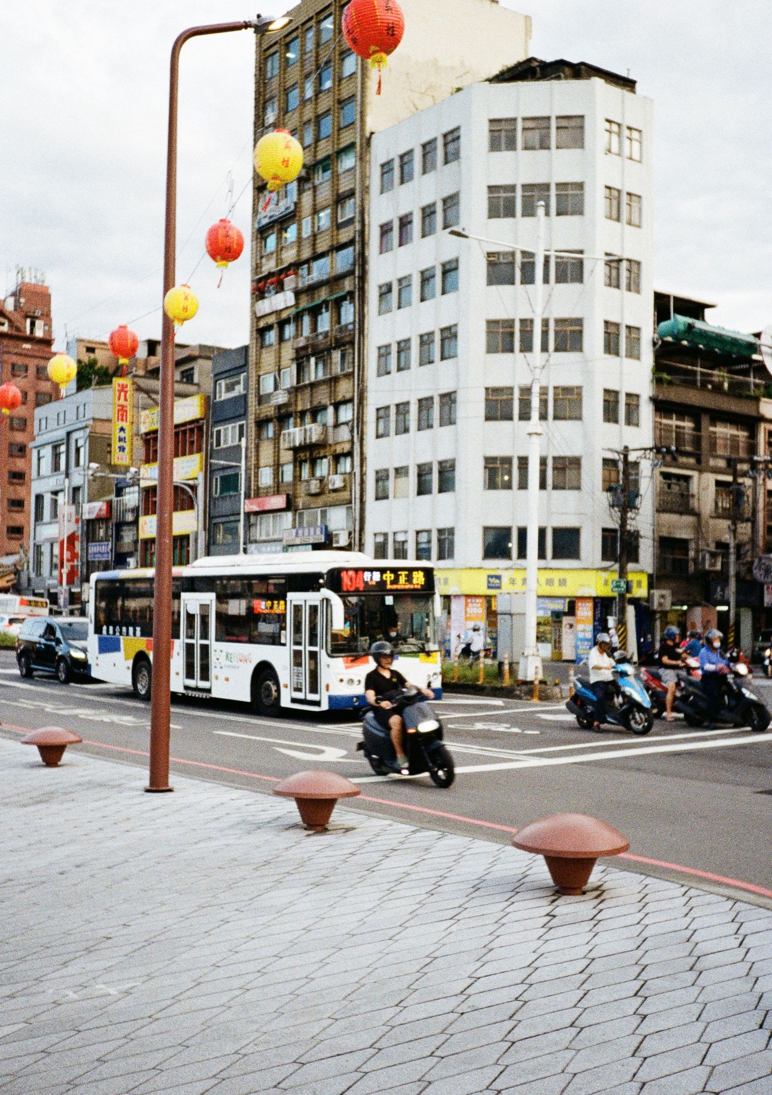Urban street corner with colorful paper lanterns and white modernist buildings under cloudy skies.
