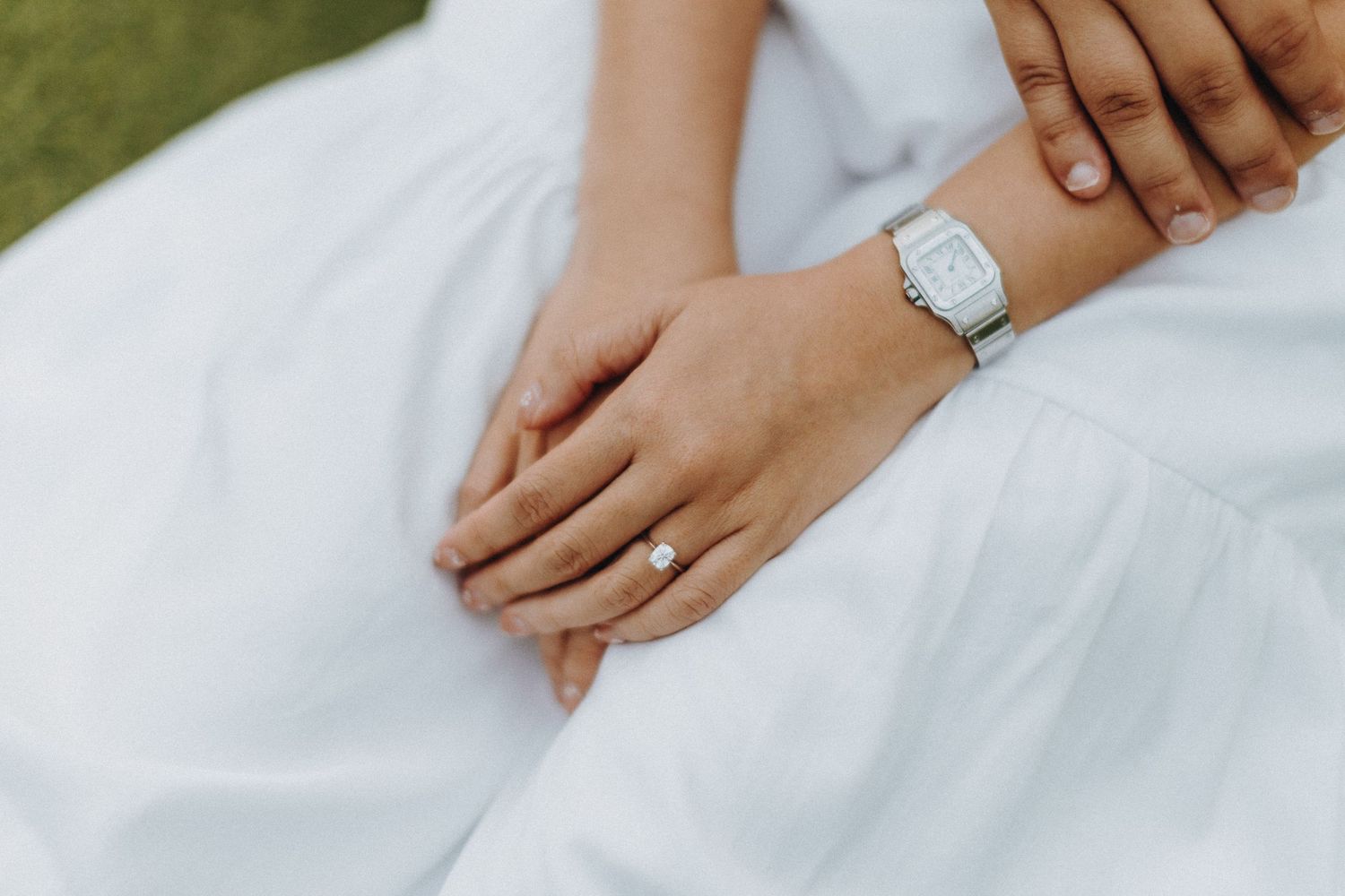 A silver wristwatch and diamond engagement ring shown on hands resting against white fabric.
