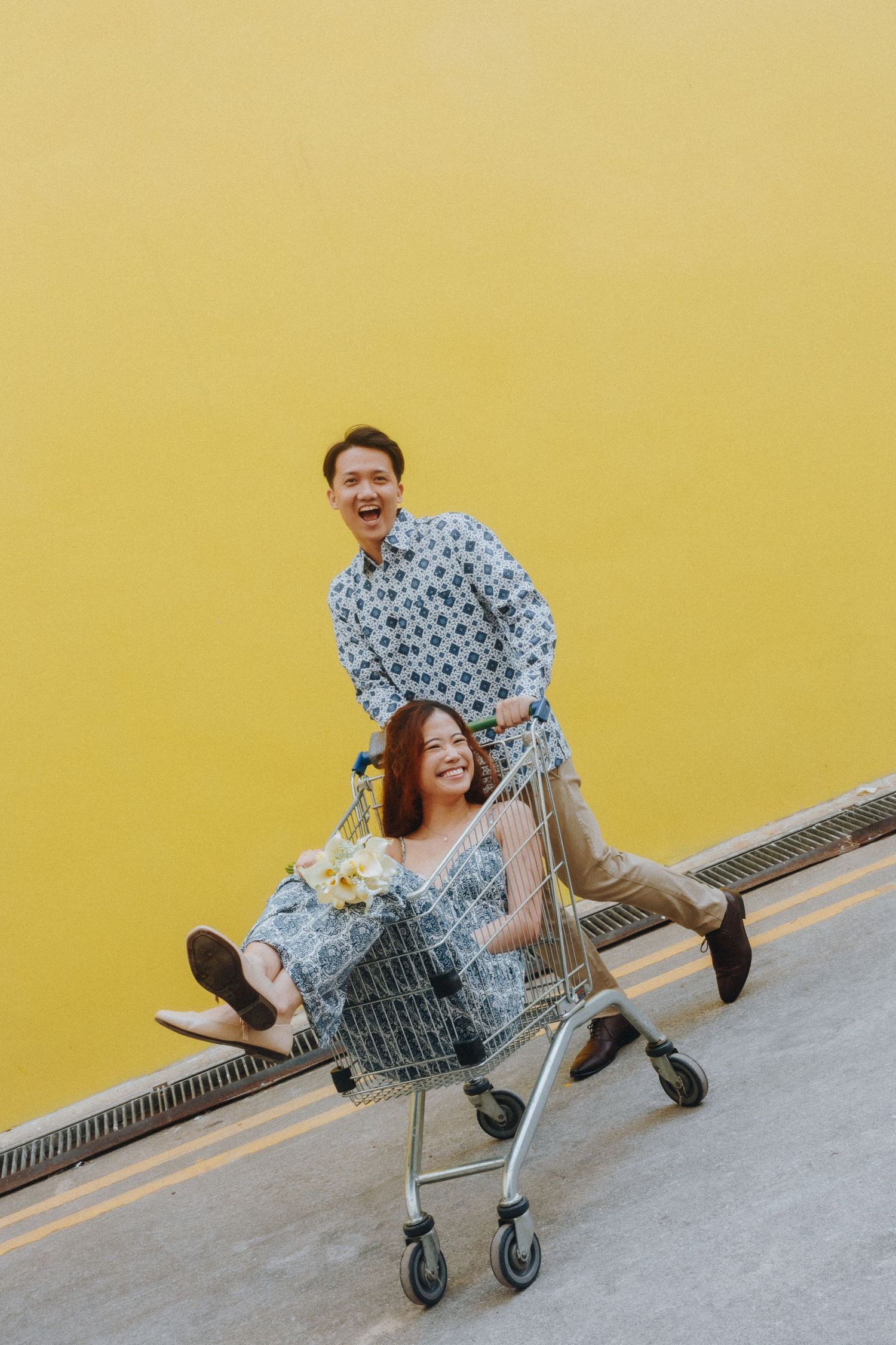 Playful couple having fun with shopping cart against yellow wall in casual outfits and smiles.