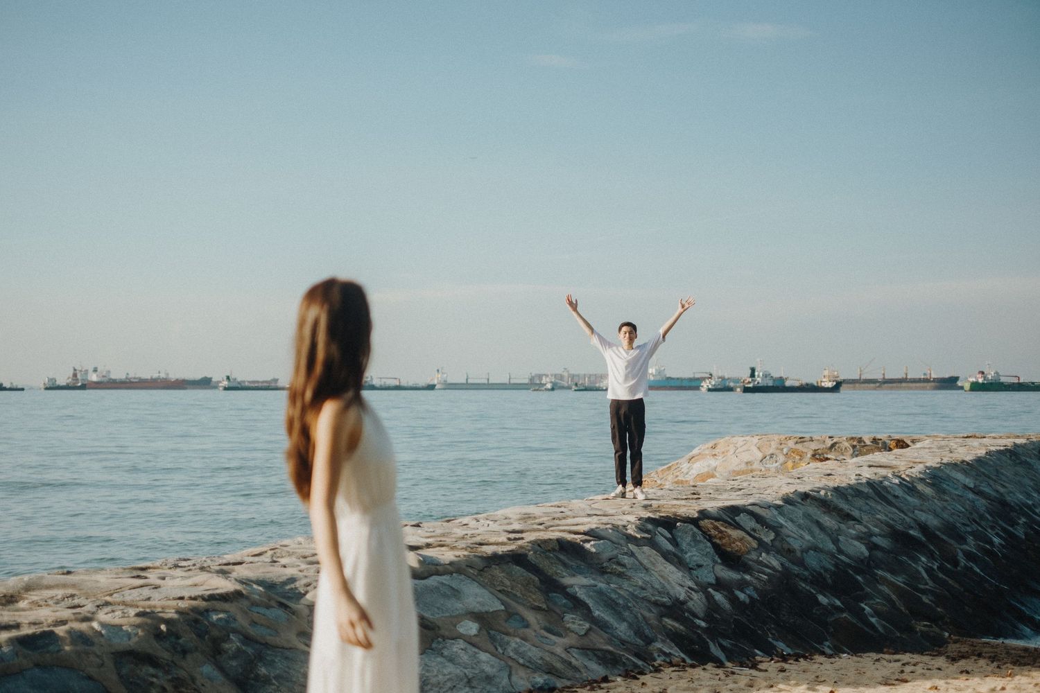 Two people share a moment on a seaside walkway with ships visible in the distance.
