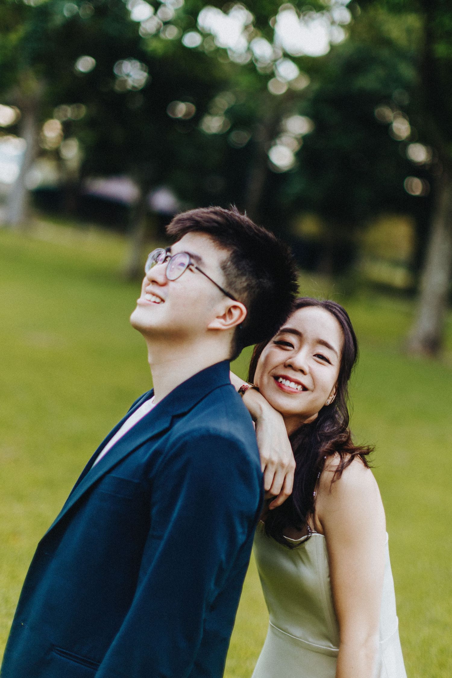 A playful couple in formal attire poses in a park with bokeh-filled background.