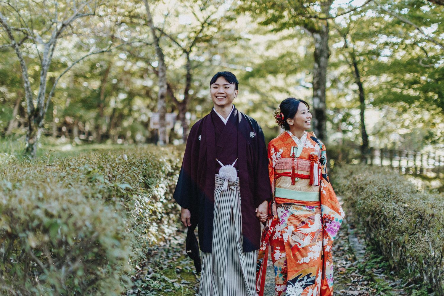 A couple in traditional kimono and western attire pose together in a serene garden setting with green foliage.