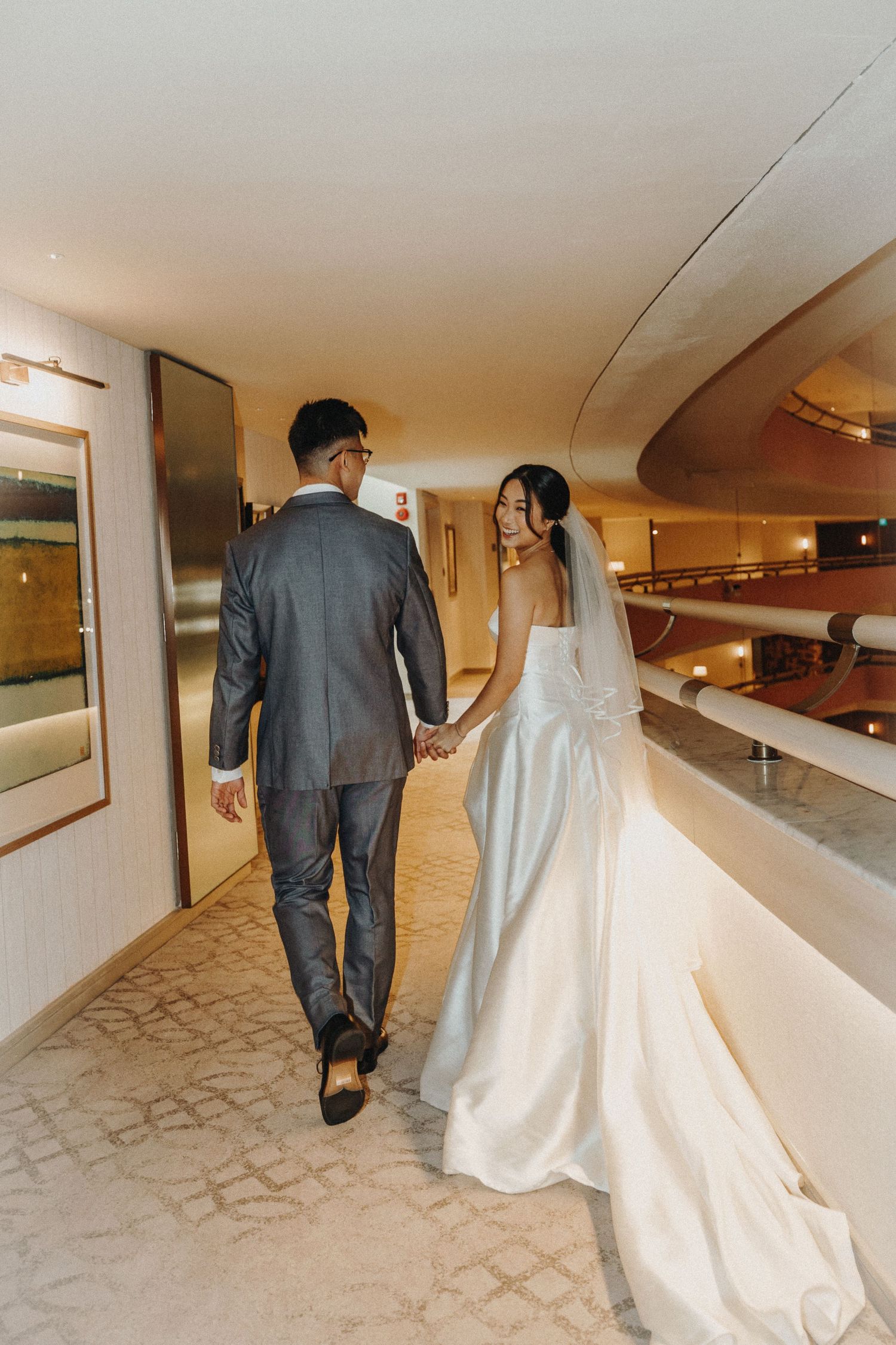 Bride in flowing white gown follows groom through a carpeted hallway.