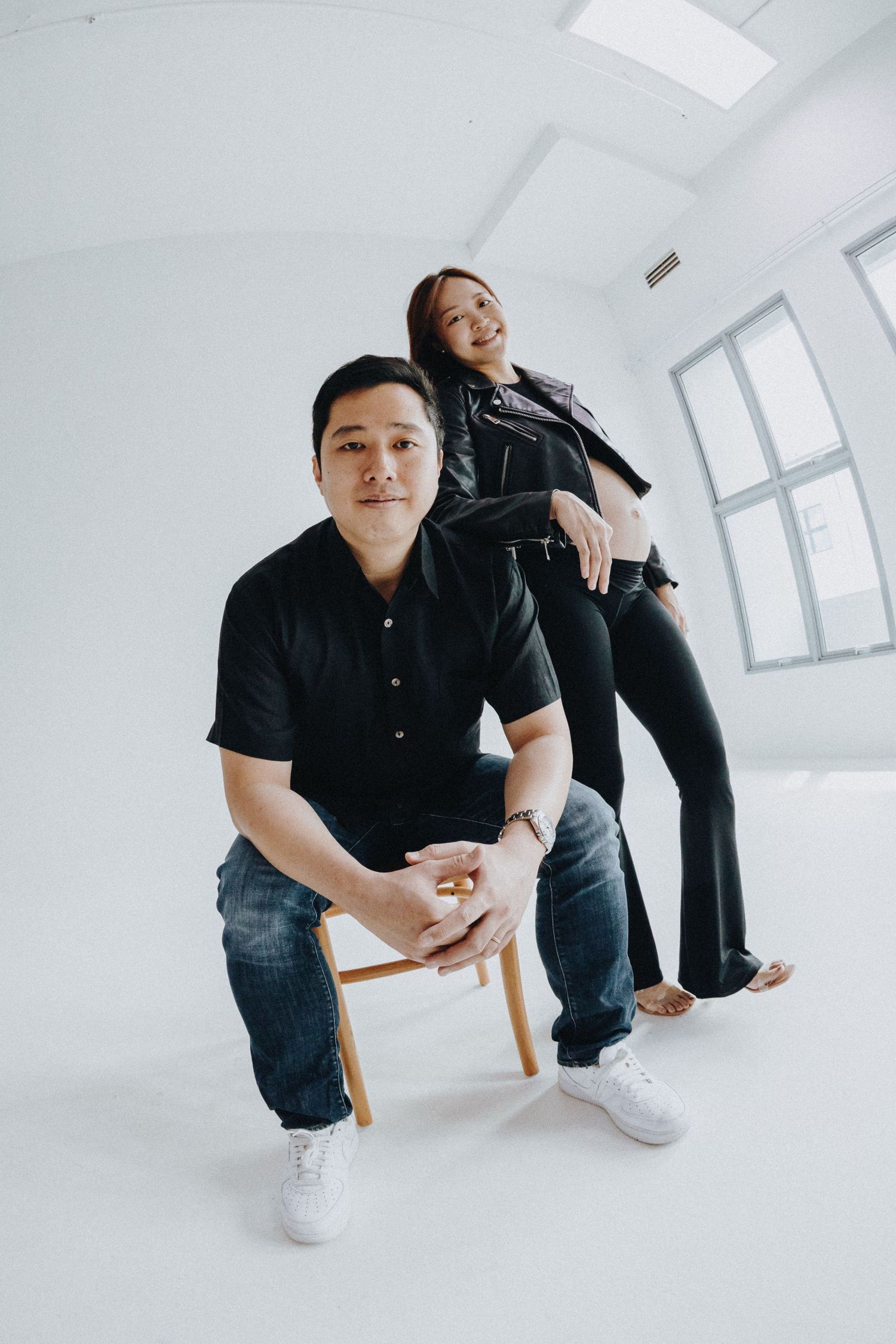 Two people in black outfits pose casually in a bright white studio setting.
