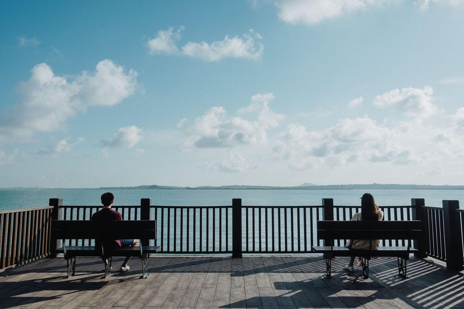 Two benches on a wooden deck overlook a calm ocean vista under puffy white clouds.