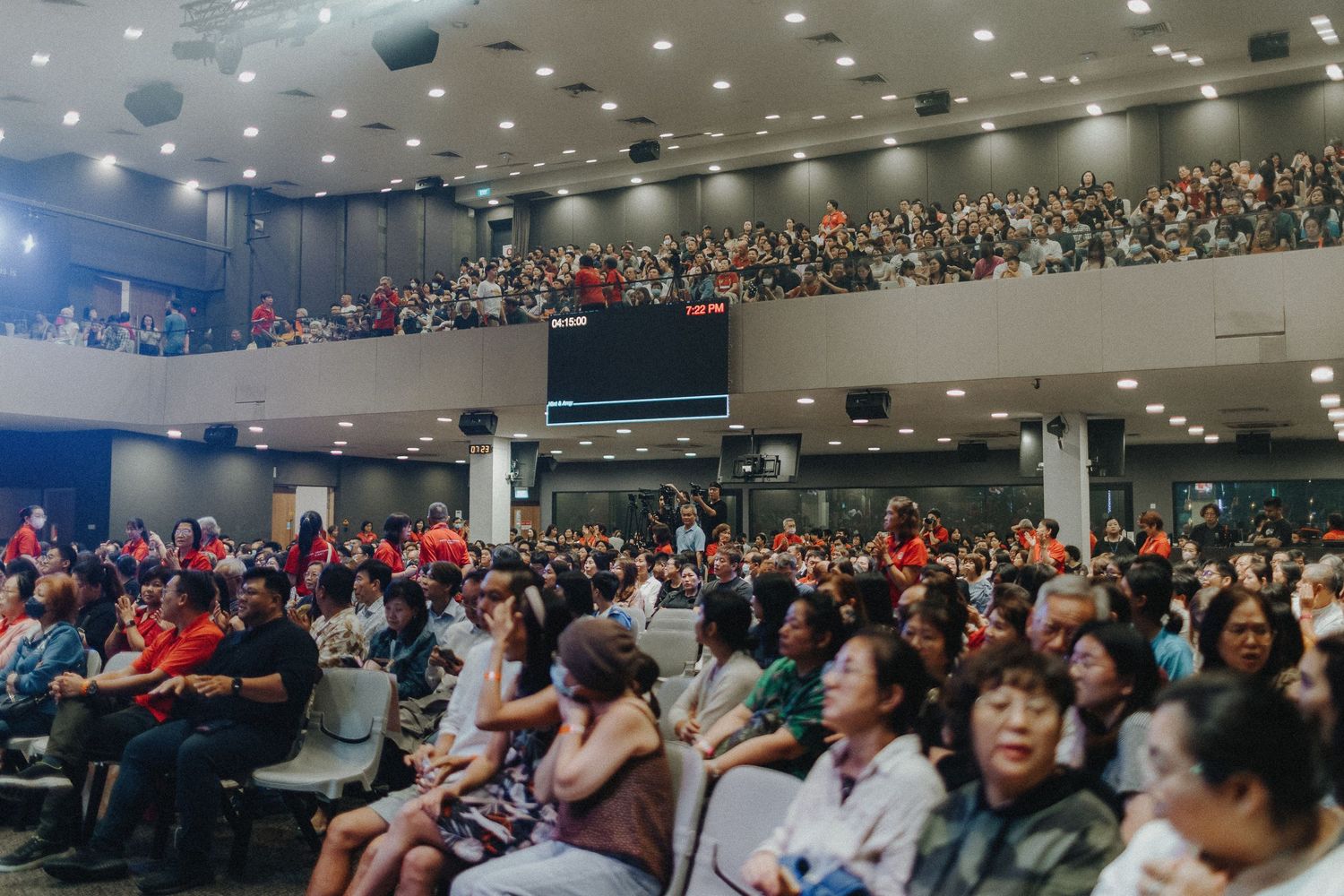 Large audience fills multiple levels of indoor auditorium seating during a crowded conference or presentation event.