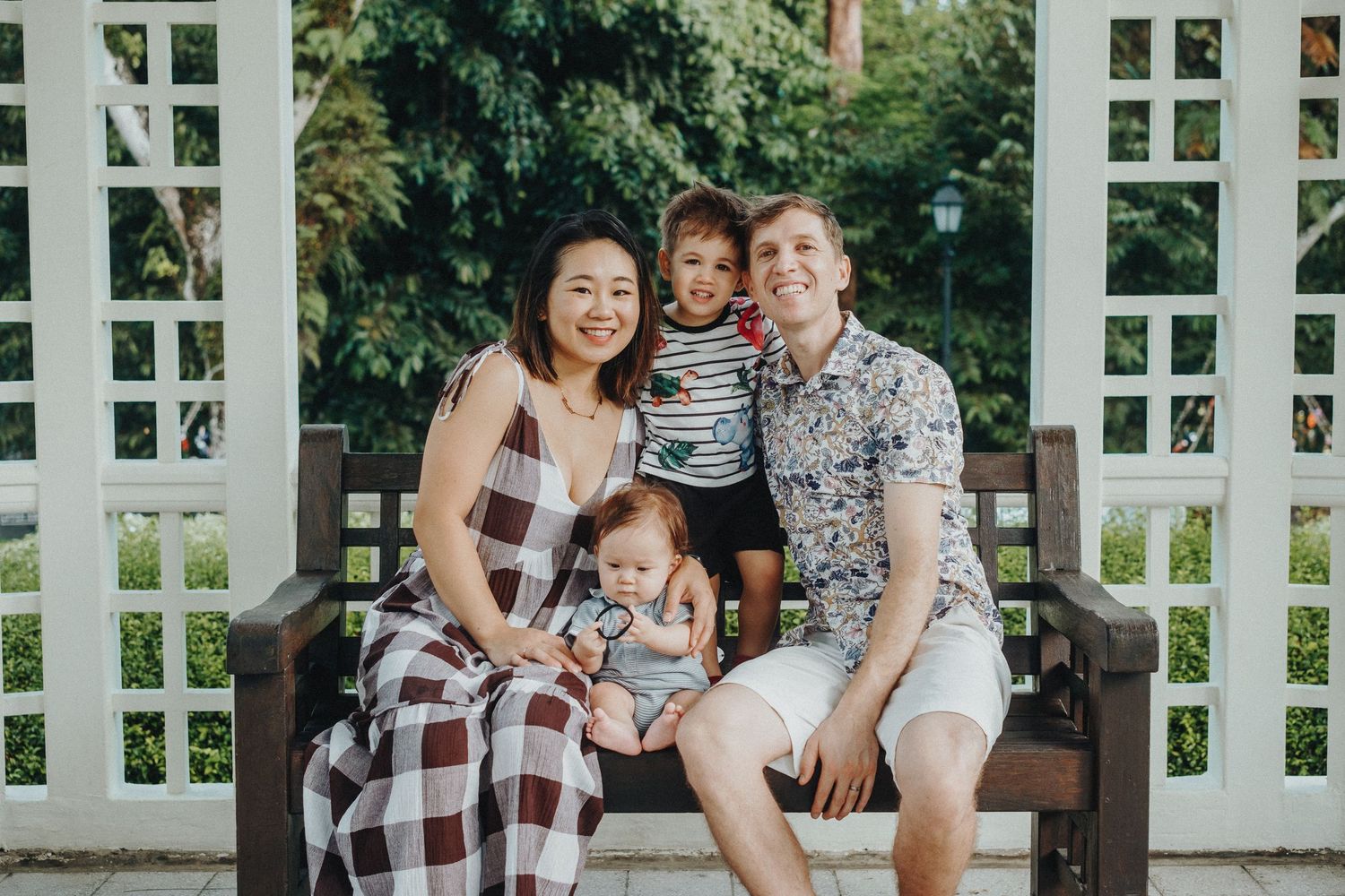 A family wearing coordinated casual outfits sits together on a wooden bench under a white garden arbor on a sunny day.
