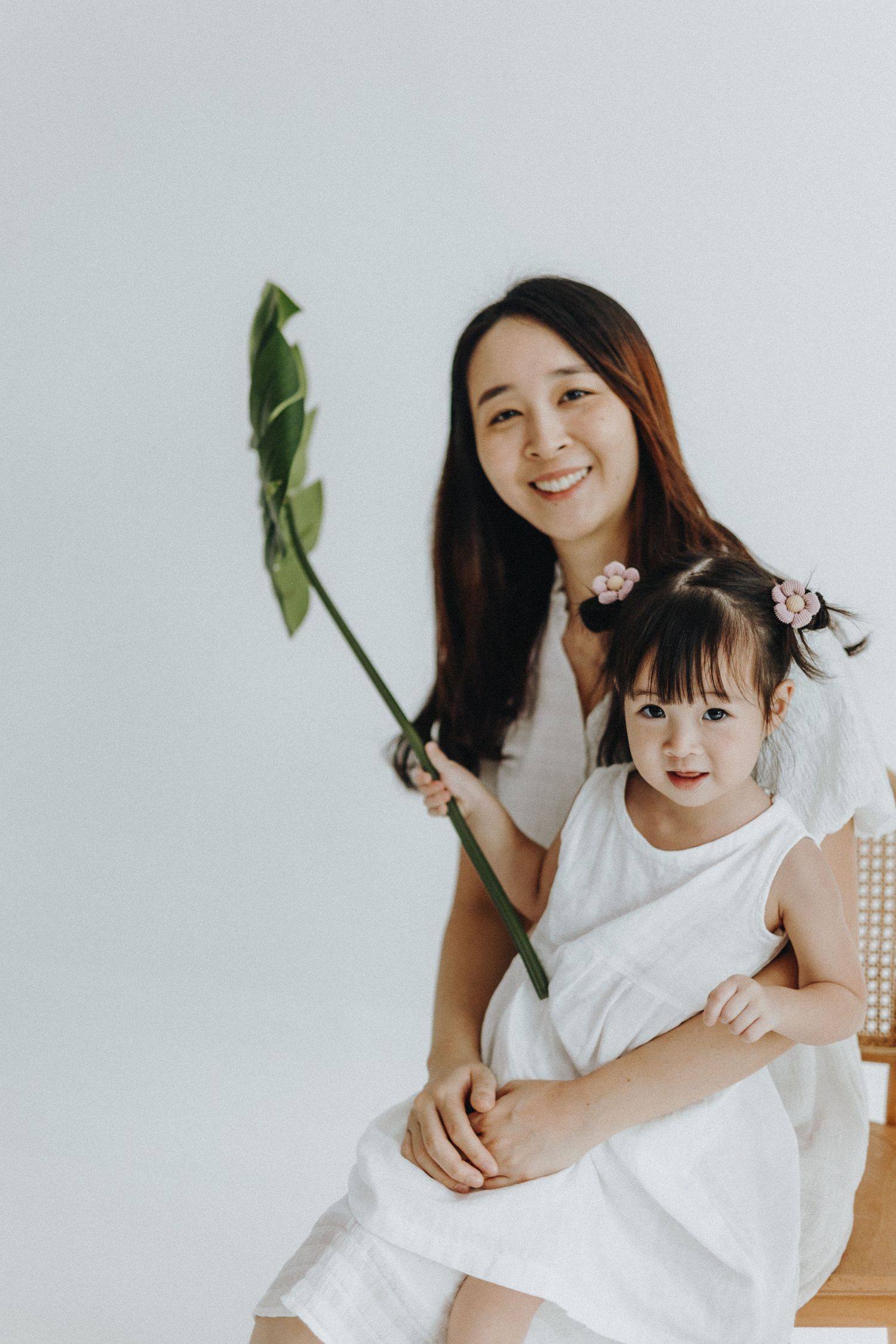 Mother and child in white outfits play with a large green tropical leaf against a light background.