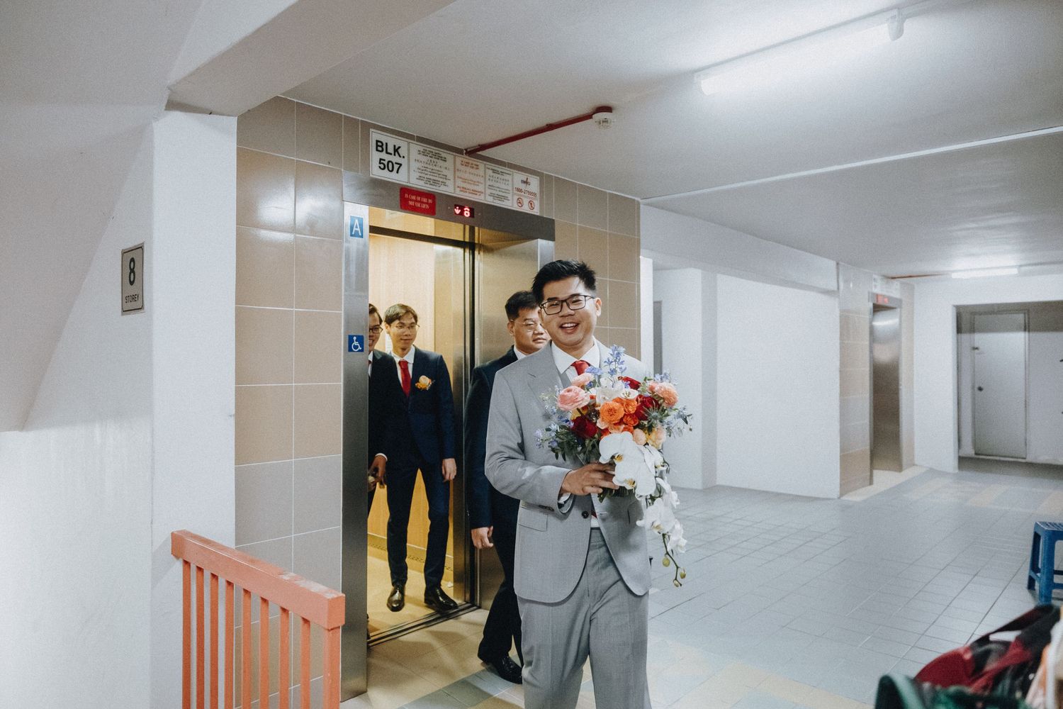 In a hallway corridor near an elevator, someone in a gray suit holds a vibrant bouquet of flowers.