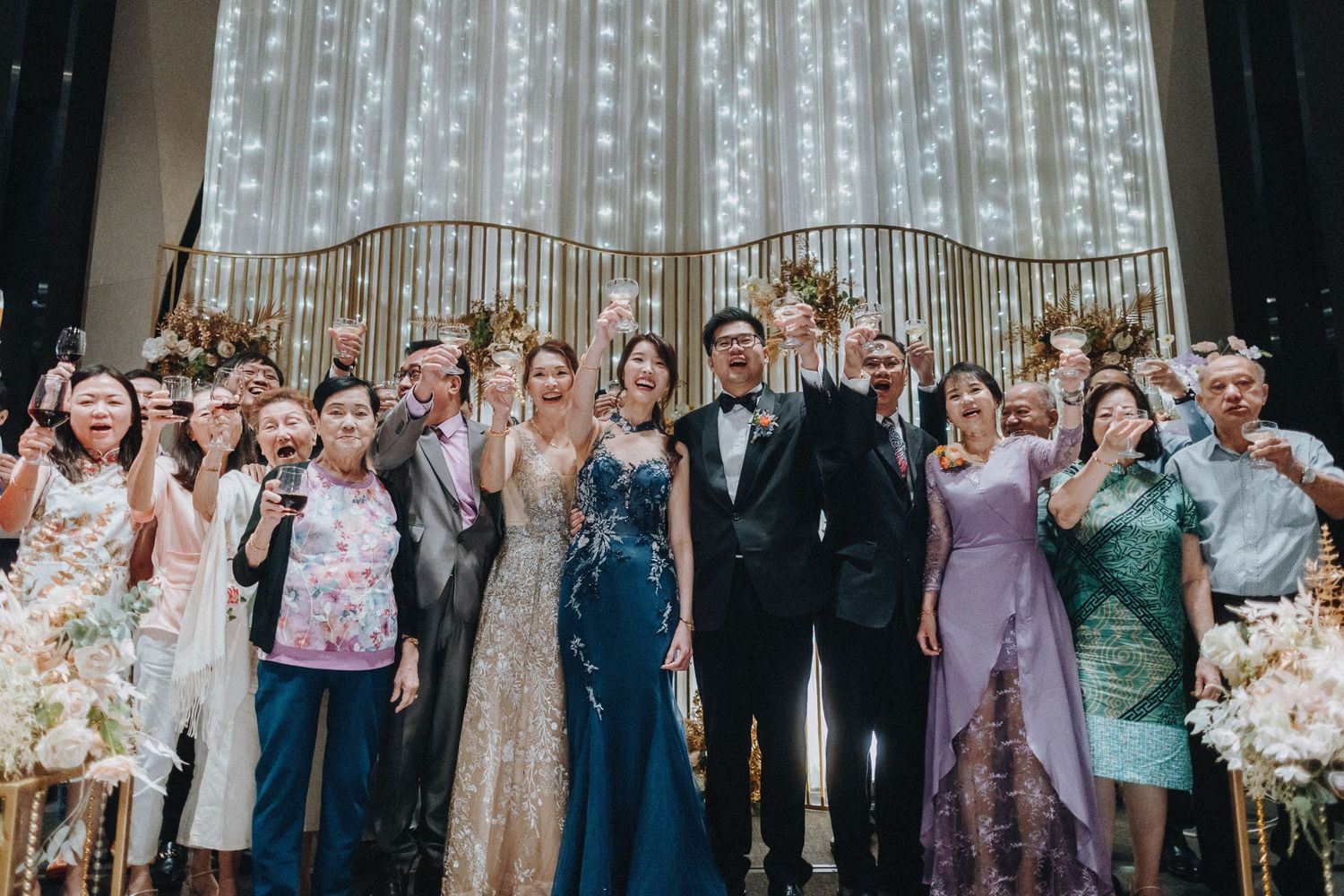 A large wedding party poses together in front of a sparkly backdrop with decorative floral arrangements.
