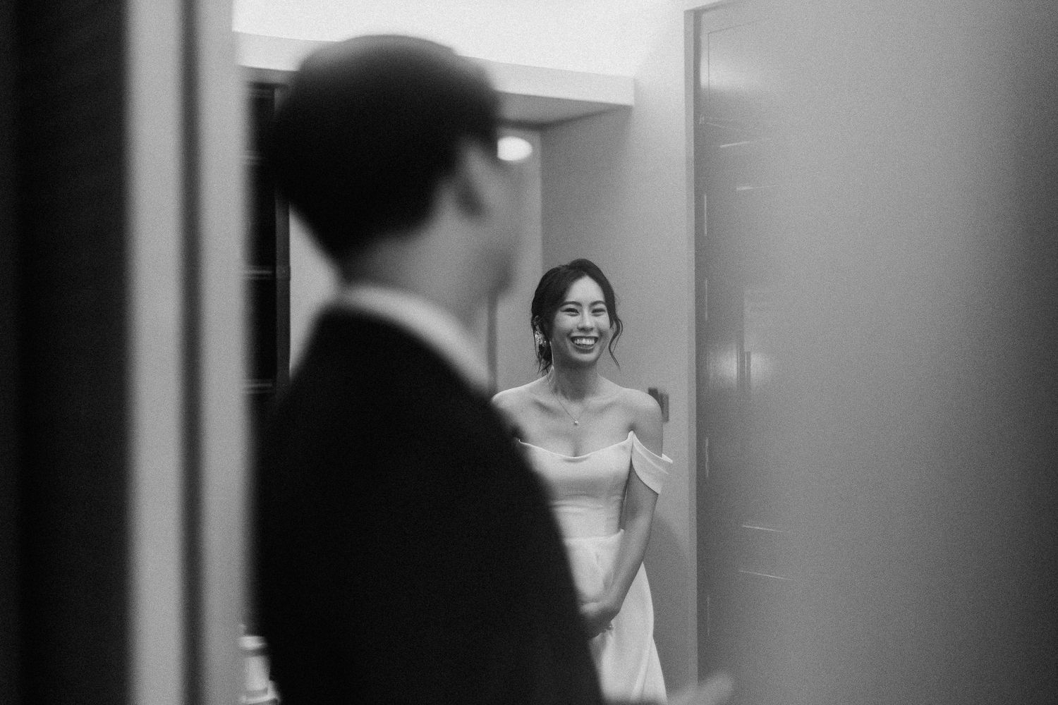 A black and white photograph captures a joyful moment during a wedding as a bride and groom share a glance through a doorway.