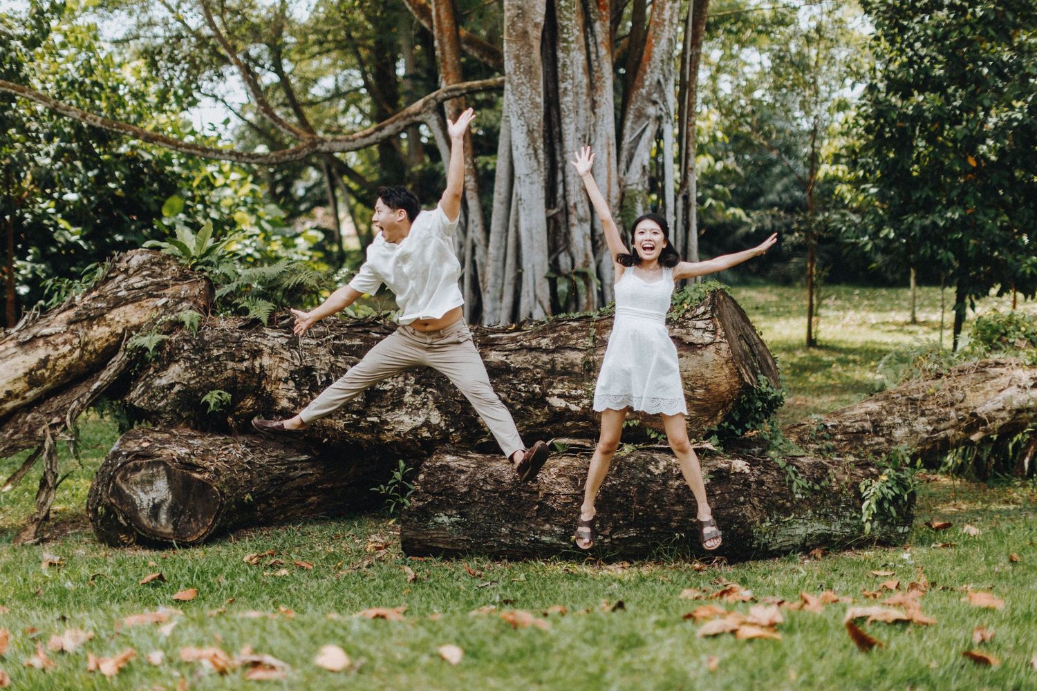 A couple strikes playful jumping poses in front of a large tree trunk outdoors.