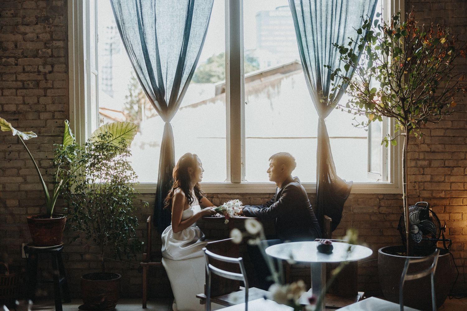Two people share an intimate conversation while sitting at a cafe table near large windows with linen curtains and brick walls.