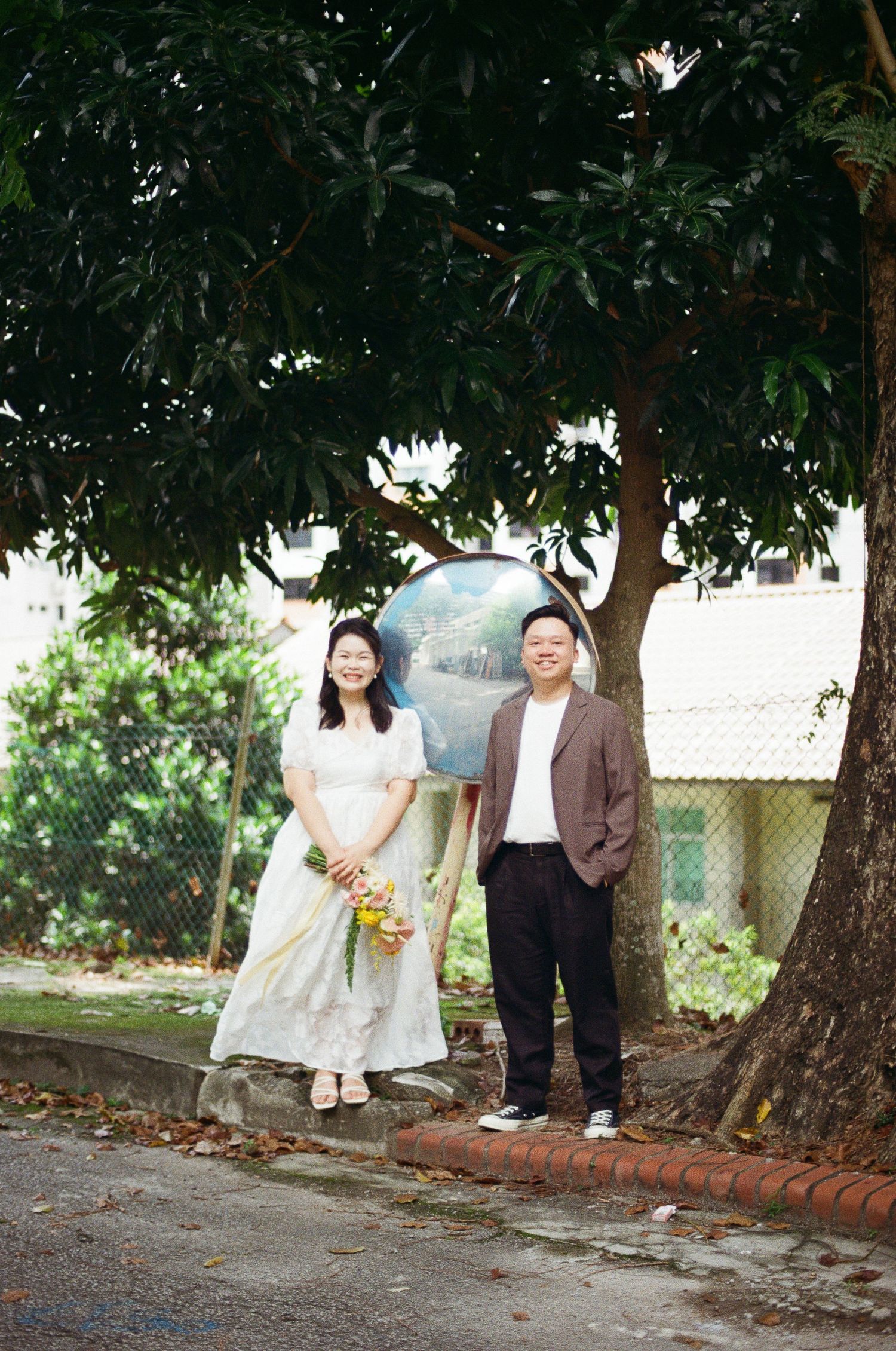 A couple poses for a romantic photo under a large tree on a sidewalk.