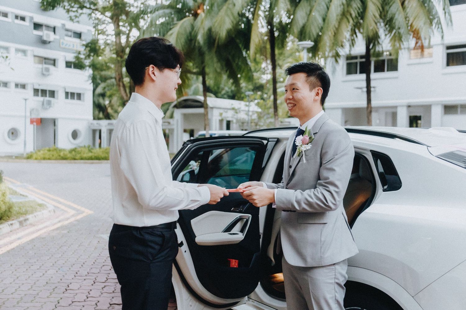 Two men in formal attire stand by a car on a palm-lined driveway during a wedding day.
