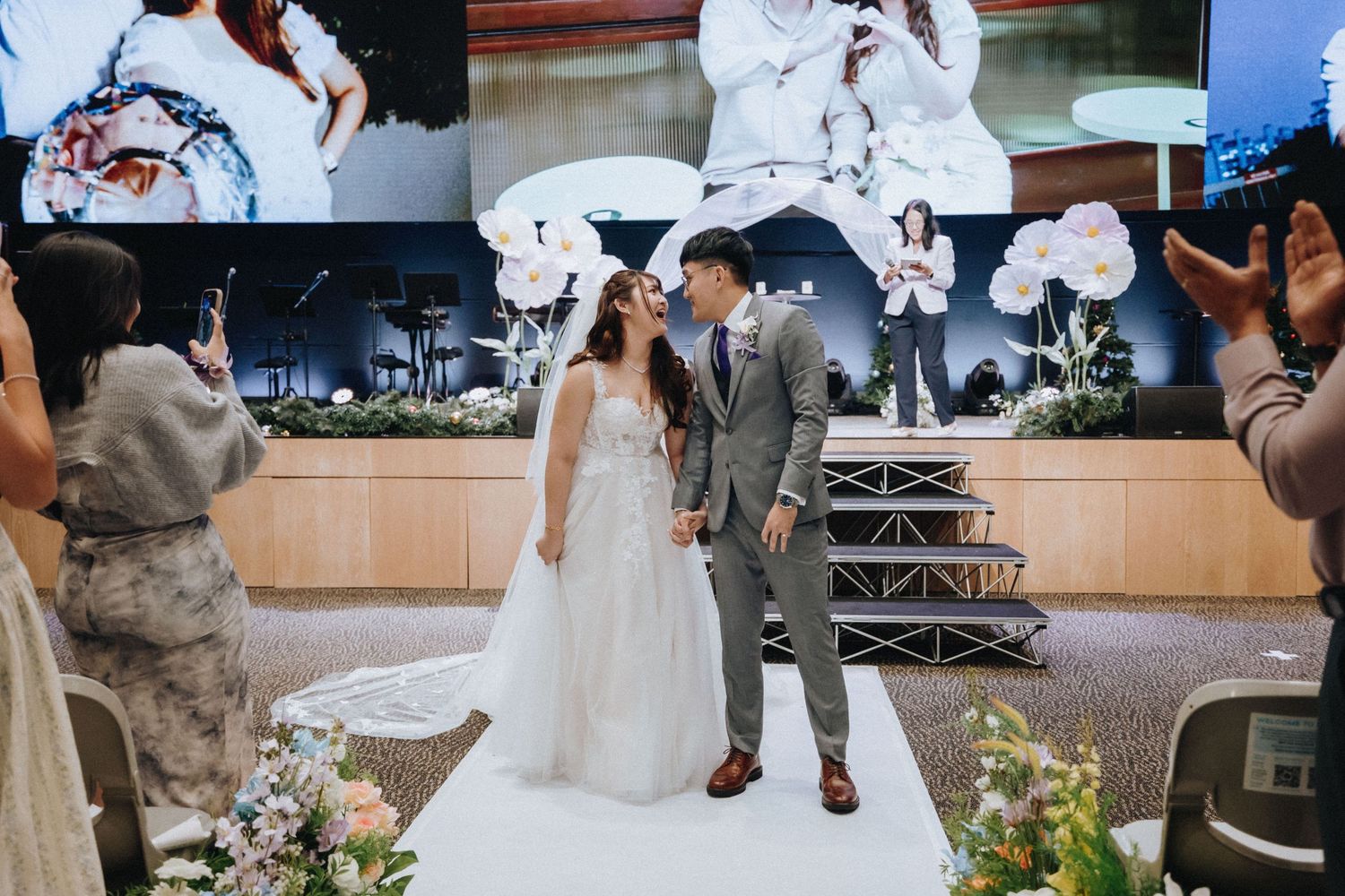 Newlyweds pose on decorated wedding stage with white floral arrangements.