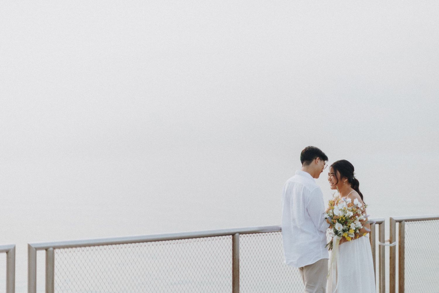 A couple in white attire stands on a balcony overlooking a misty view while holding a floral bouquet.
