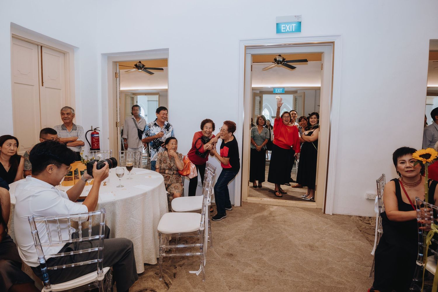 Guests mingling and celebrating at an indoor event venue with white tables, chairs, and open doorways.
