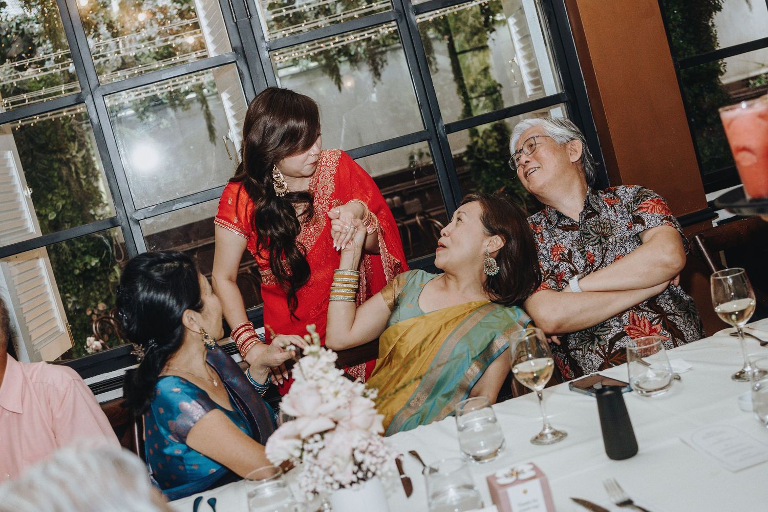 A bride in red laughs with female family members seated at a wedding reception table.