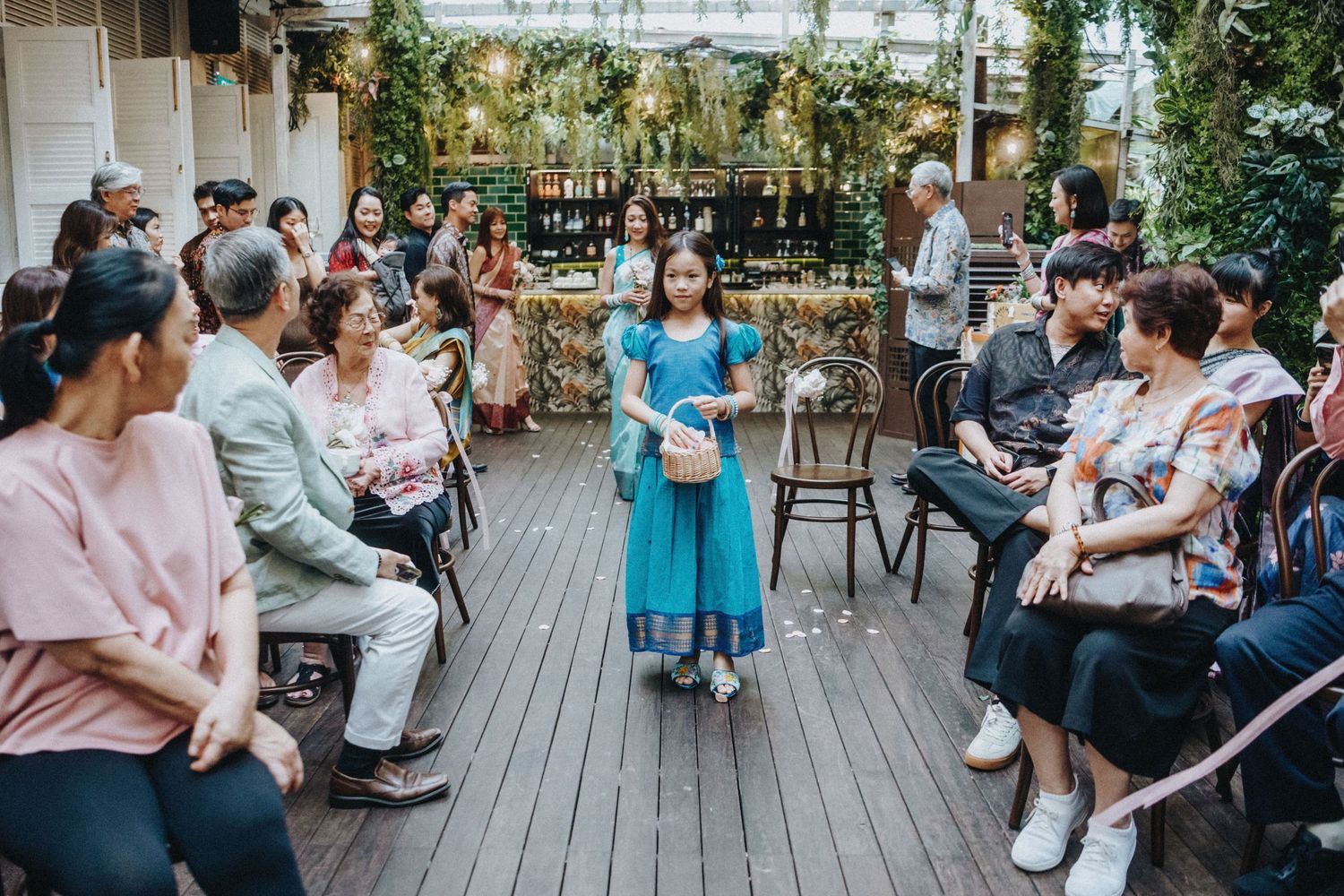 Young girl in blue dress walks through seated guests at an outdoor Asian wedding celebration on a wooden deck.