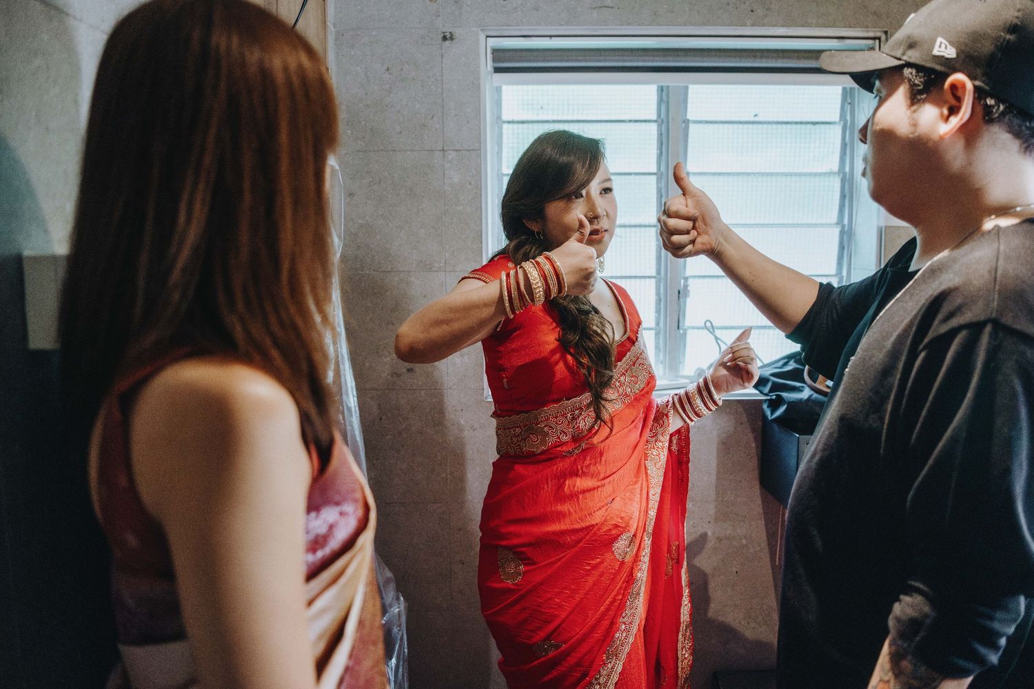 Woman in red saree with bangles gestures while talking to two people indoors during a traditional Indian celebration.