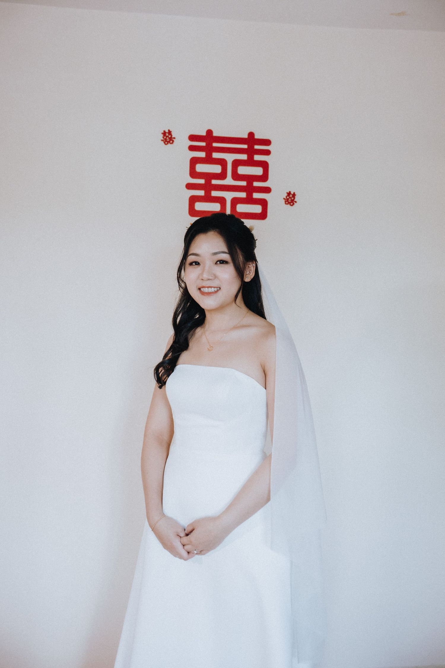 Smiling Asian bride in strapless white gown and veil poses before white wall with red double happiness symbol.