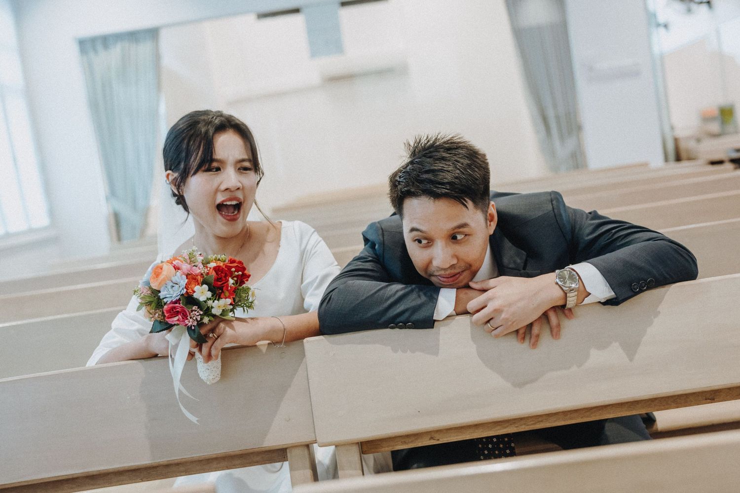 Joyful newlywed couple posing playfully in church pews, bride holding red bouquet in white dress.