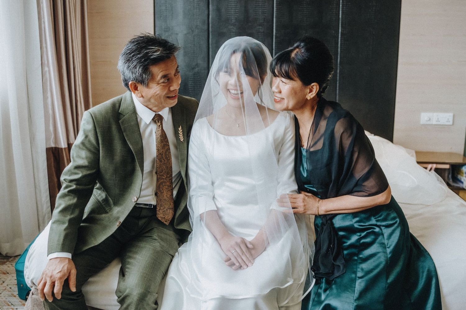 Bride in white gown and veil sits with parents on bed in hotel room before wedding ceremony.