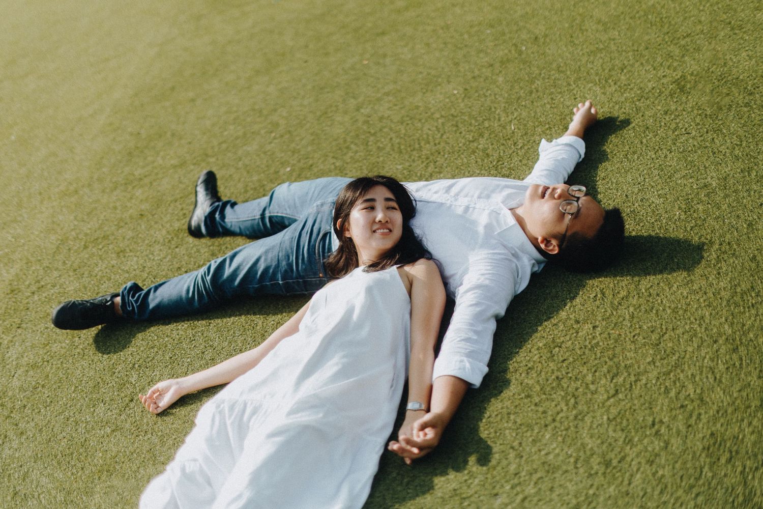 A couple lies on bright green grass wearing white and blue casual attire.
