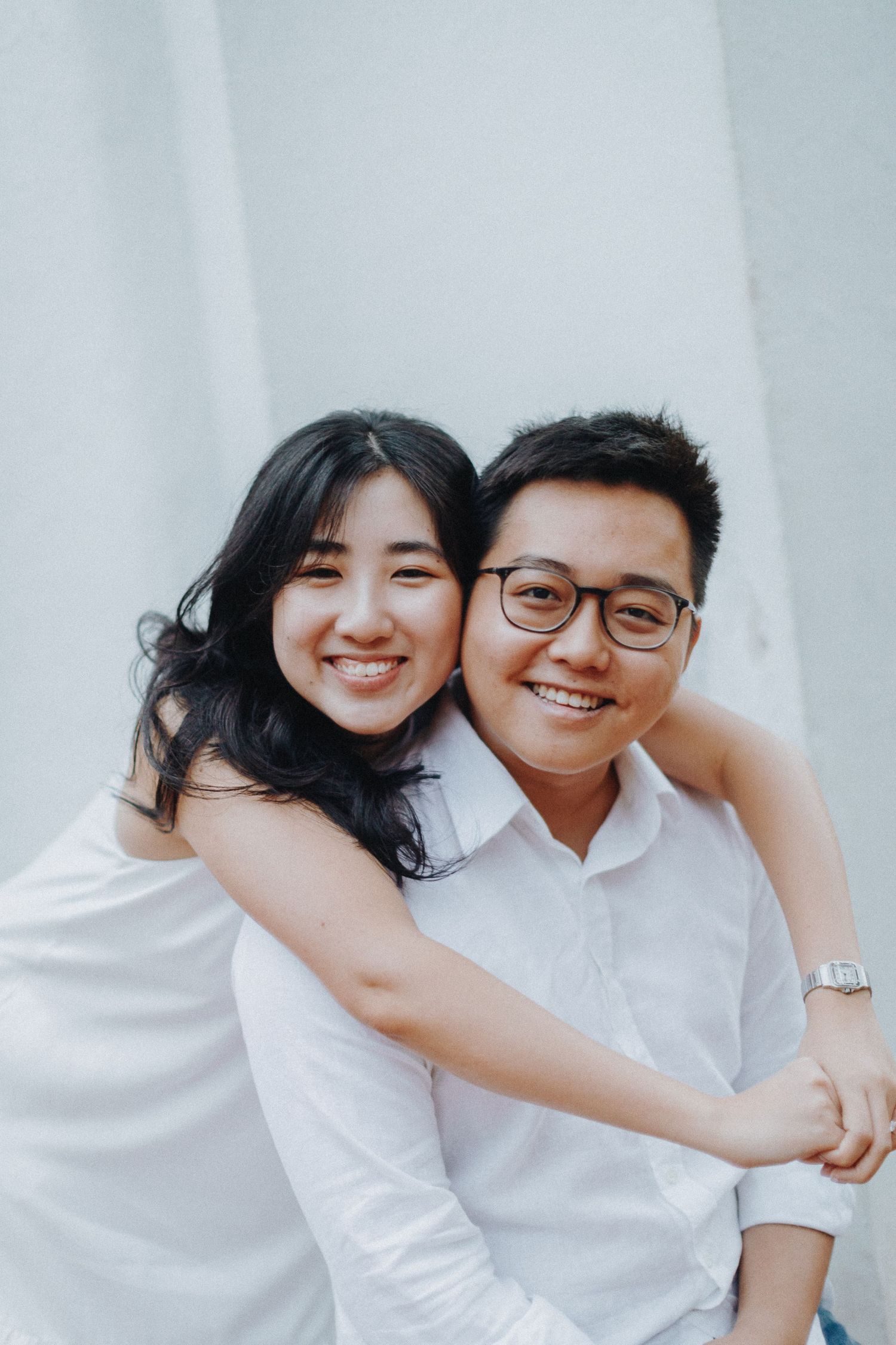 A couple wearing white outfits embrace and smile warmly against a light background.