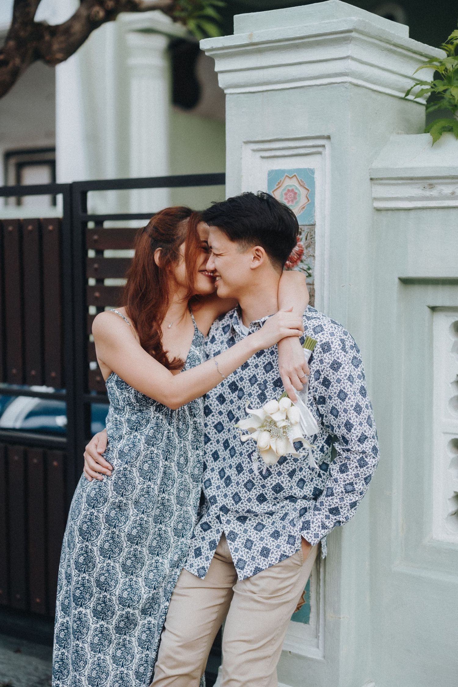 A romantic couple in patterned clothing embraces and shares a tender moment against a white pillar outdoors.