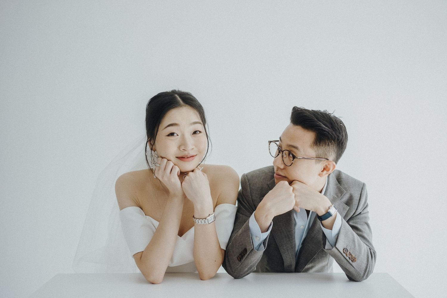 A couple in wedding attire pose playfully with their chins resting on their hands against a white backdrop.