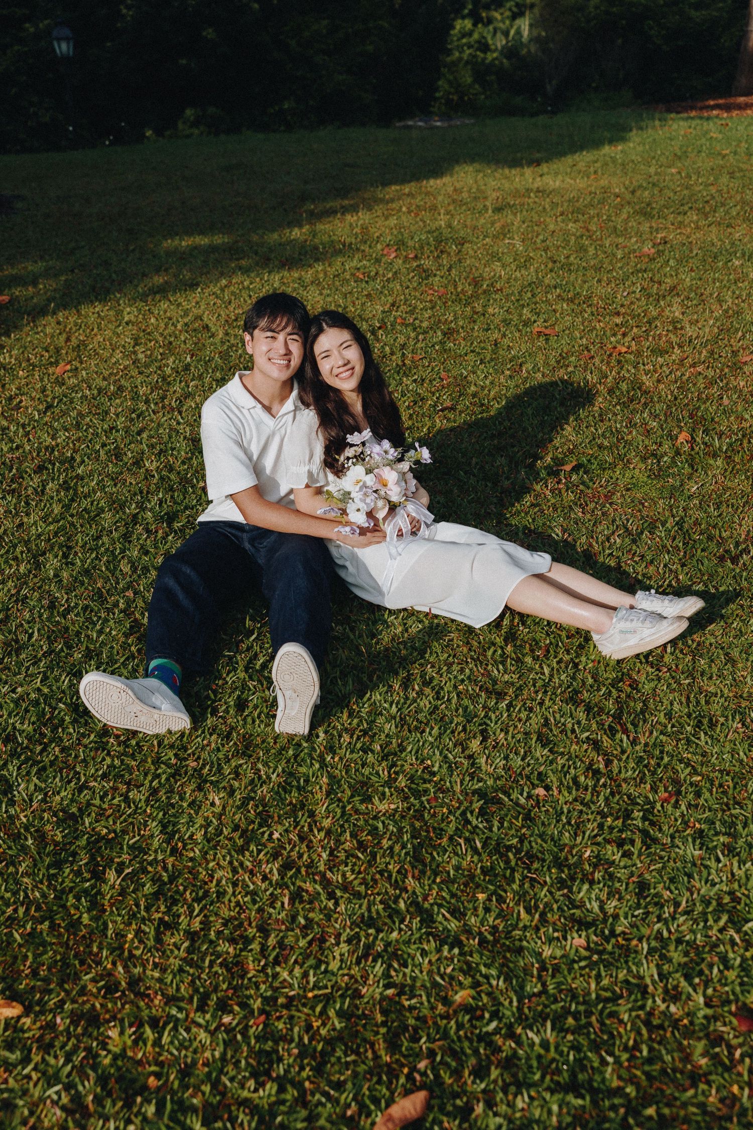 Couple lounging on grass with bouquet during outdoor photoshoot.