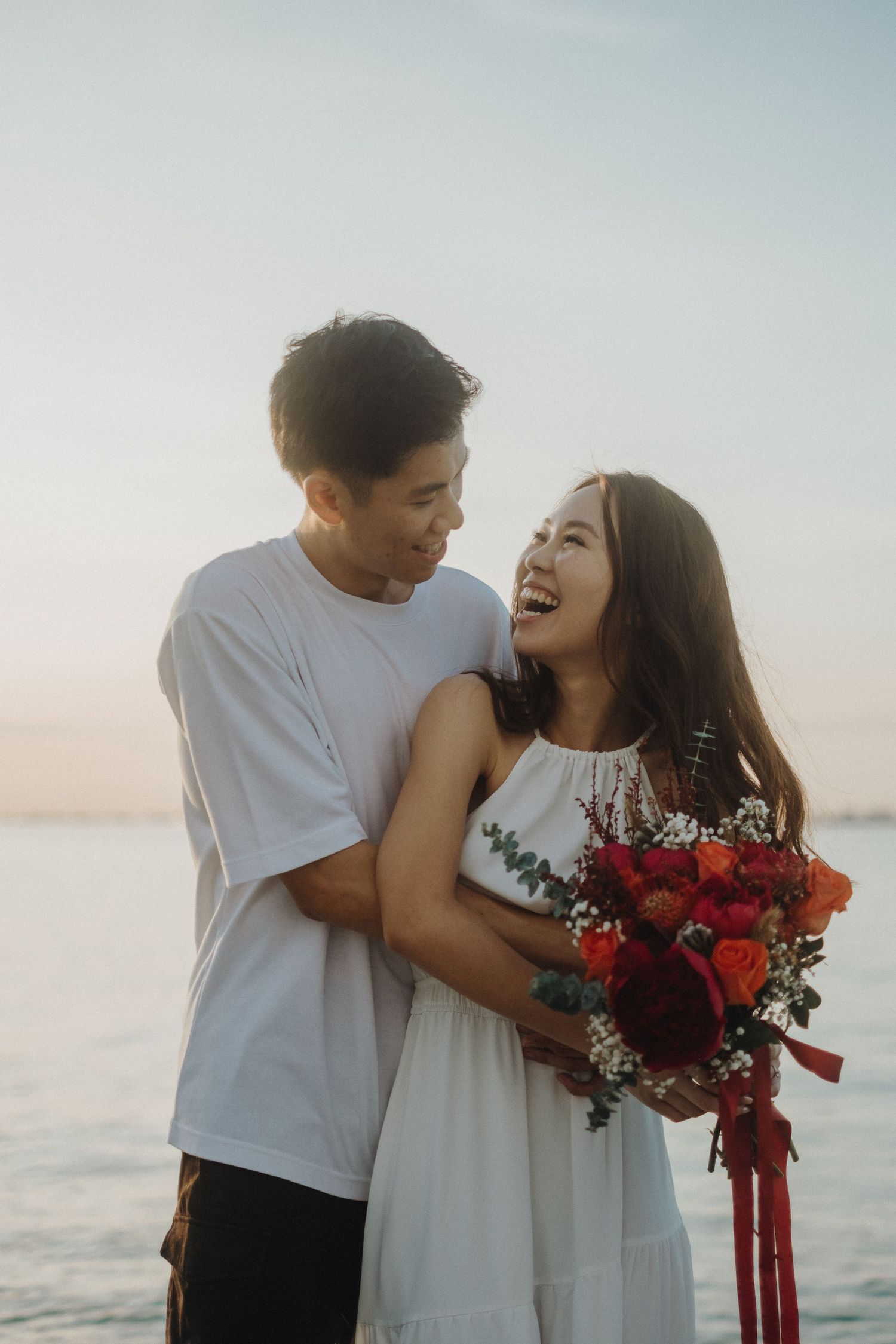 A romantic seaside moment as a couple embraces and laughs while holding a vibrant red bouquet at sunset.